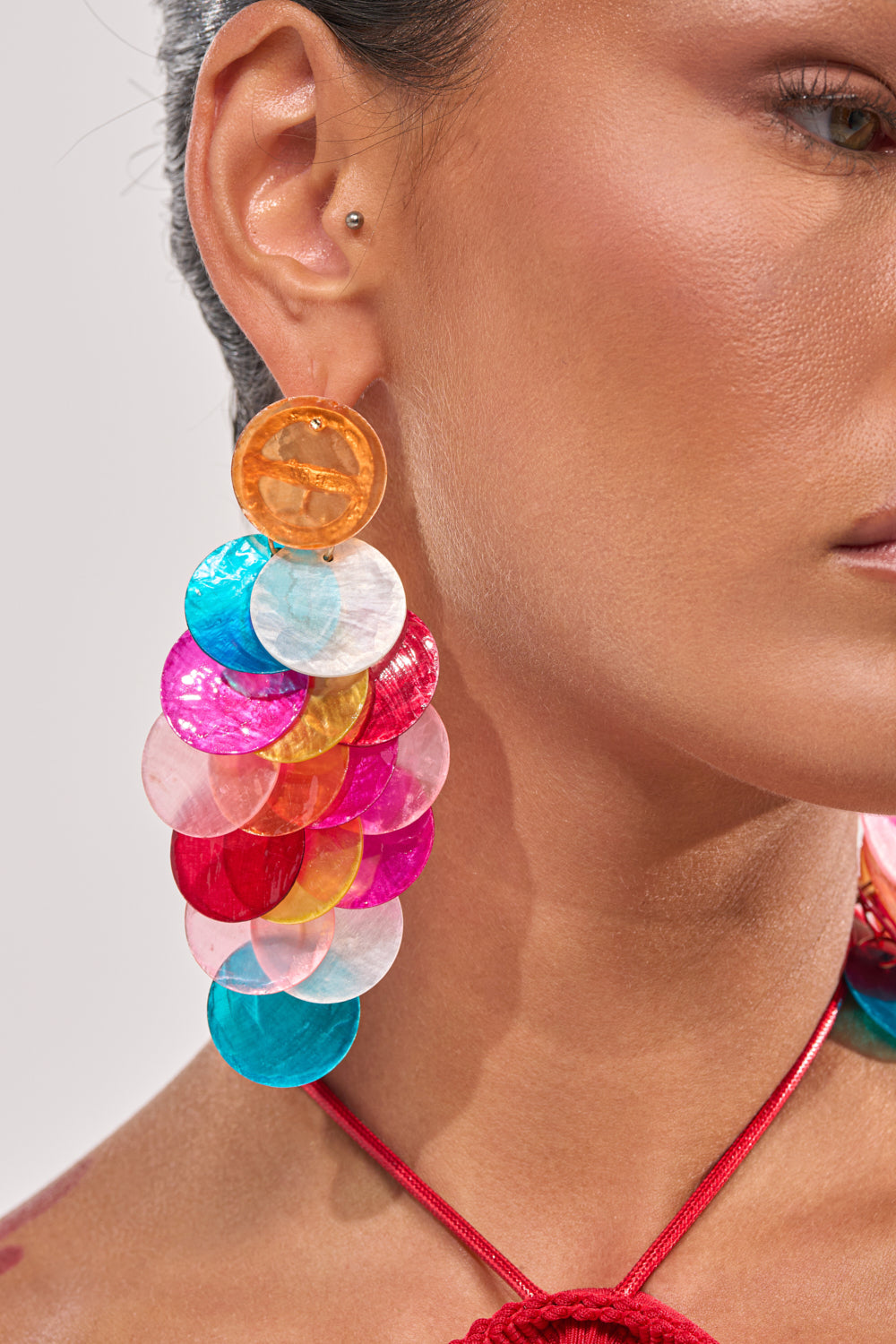 Close-up of a person wearing IM THE PARTY EARRING, featuring large multicolor overlapping circular discs in orange, pink, blue, white, and gold. The statement earring shines against their tan skin and red necklace. 2