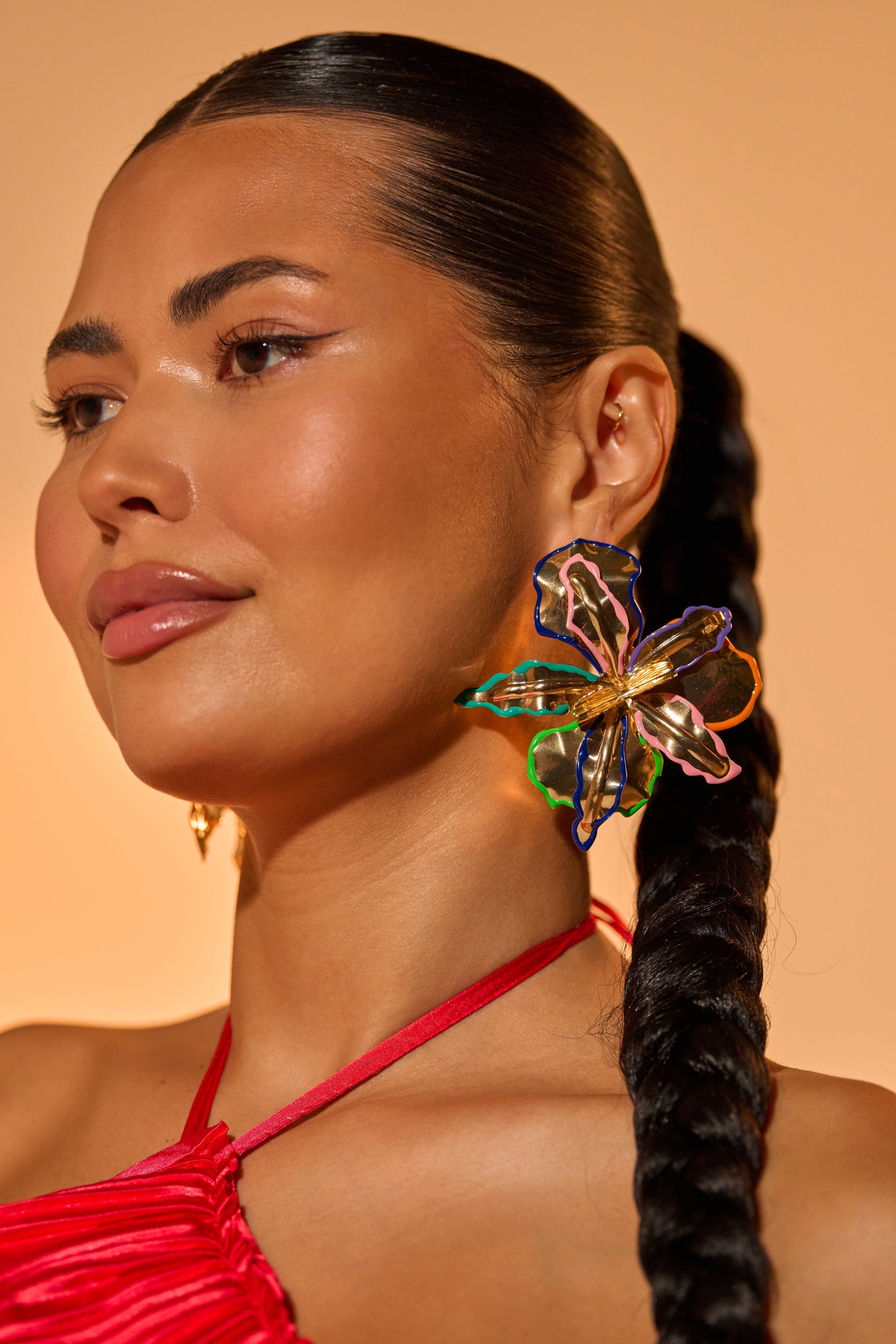 A woman with smooth, glowing skin and braided hair wears a bright red halter top and the bold, multicolor FLY ME OUT EARRING, posing against a warm beige background. 2