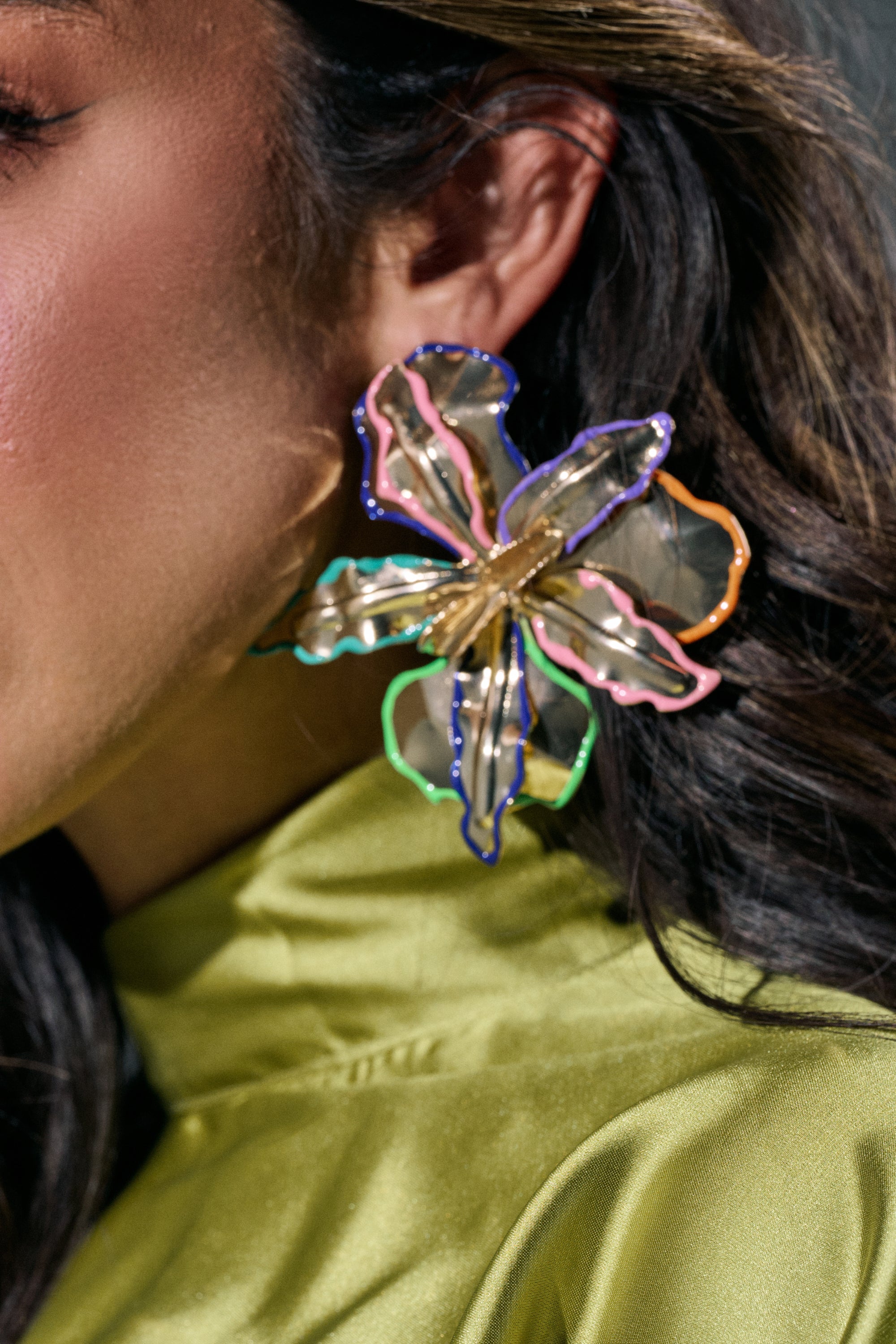 A close-up shows a person wearing the FLY ME OUT EARRING, a multicolor floral statement piece with metallic edges. The large, translucent flower-shaped earring pairs beautifully with their shiny light green top. Only part of their face and ear are visible. 1