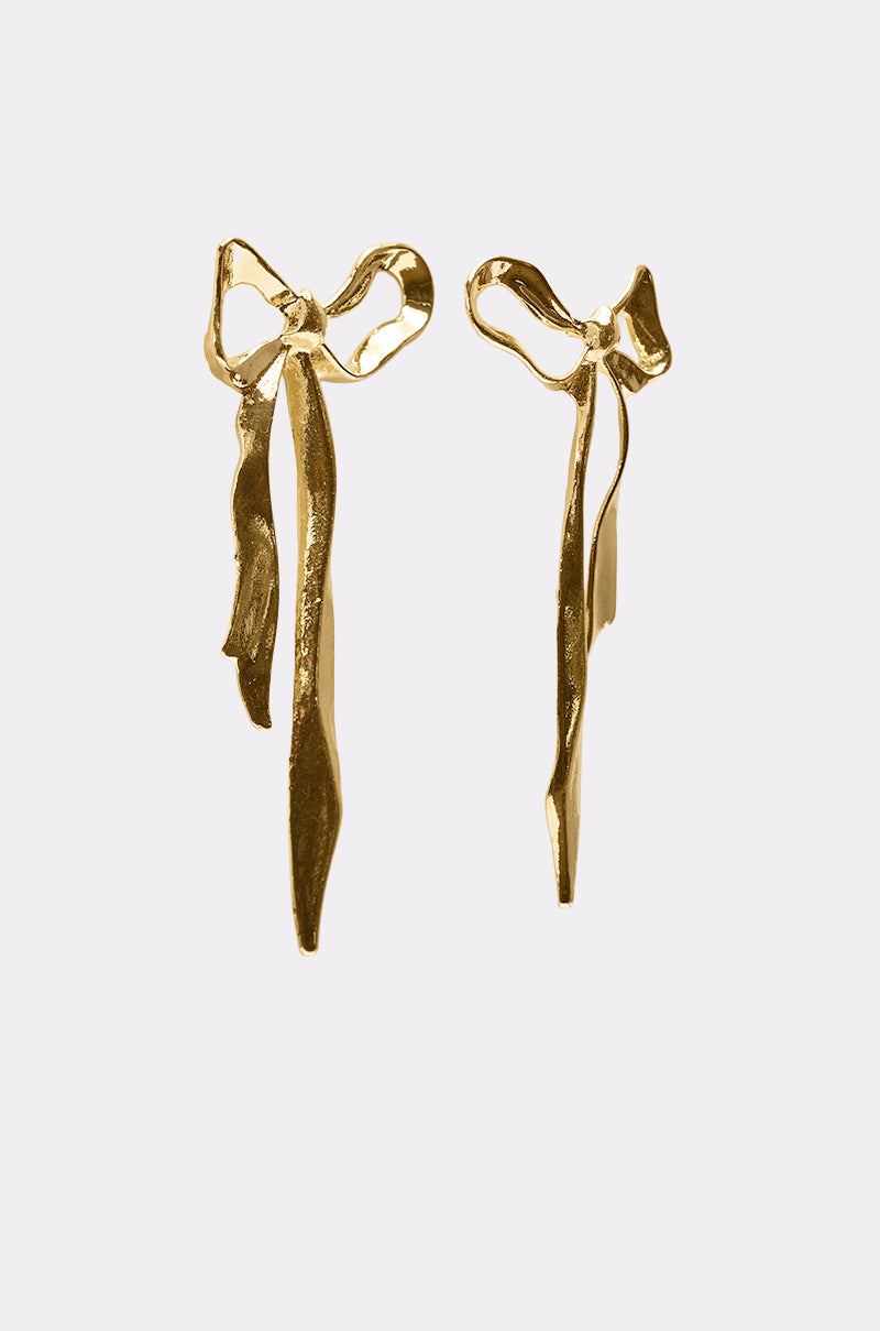 The TIE IT WITH A BOW EARRING features gold drop earrings shaped like delicate, abstract bows with long, flowing tails, displayed on a simple light background. 2