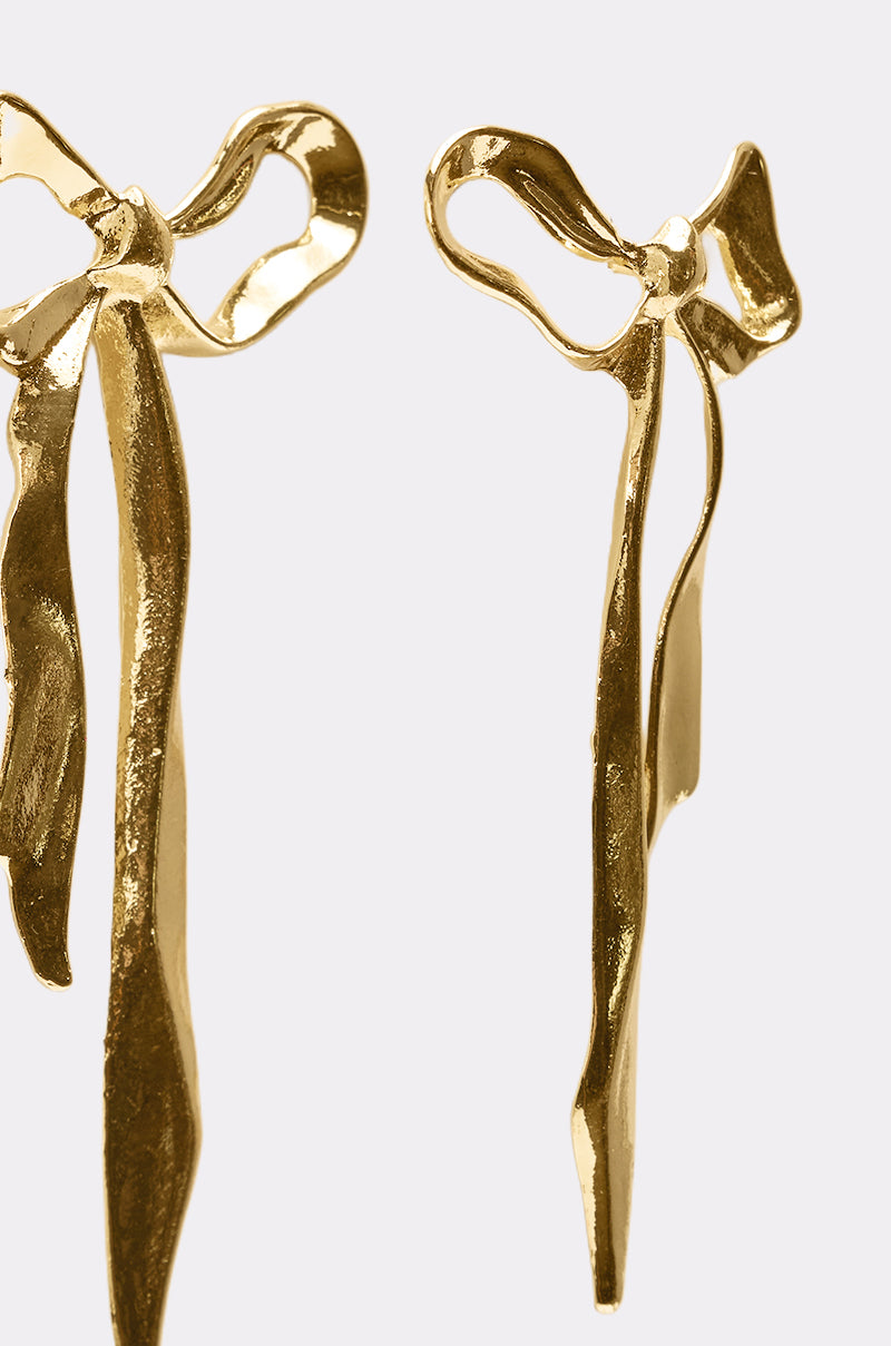 The TIE IT WITH A BOW EARRING features two shiny gold sculptural bow earrings with flowing, elongated ribbon-like tails, displayed against a plain white background. 3