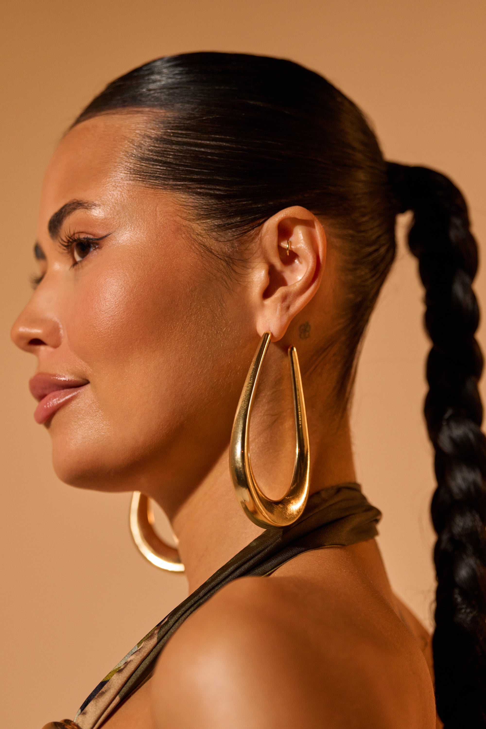 A woman with smooth, glowing skin and a long, braided ponytail models the BELLE EARRING—striking gold-tone drop earrings—shown in profile against a warm beige background. 0