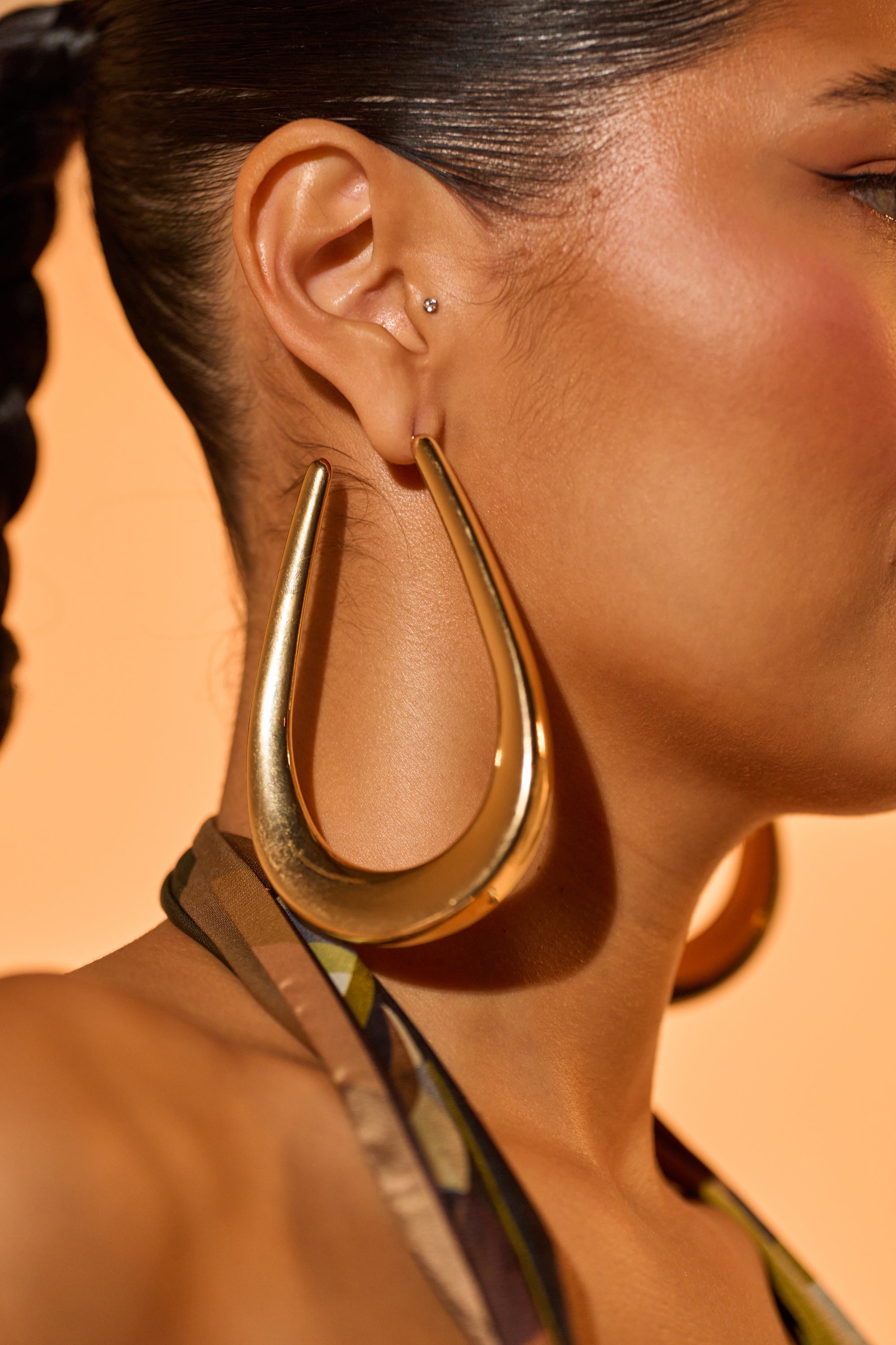 A woman with dark braided hair wears the BELLE EARRING, a large, shiny gold-tone teardrop hoop. A small stud sits in her upper ear, and a warm peach background highlights the elegant drop earring. 1