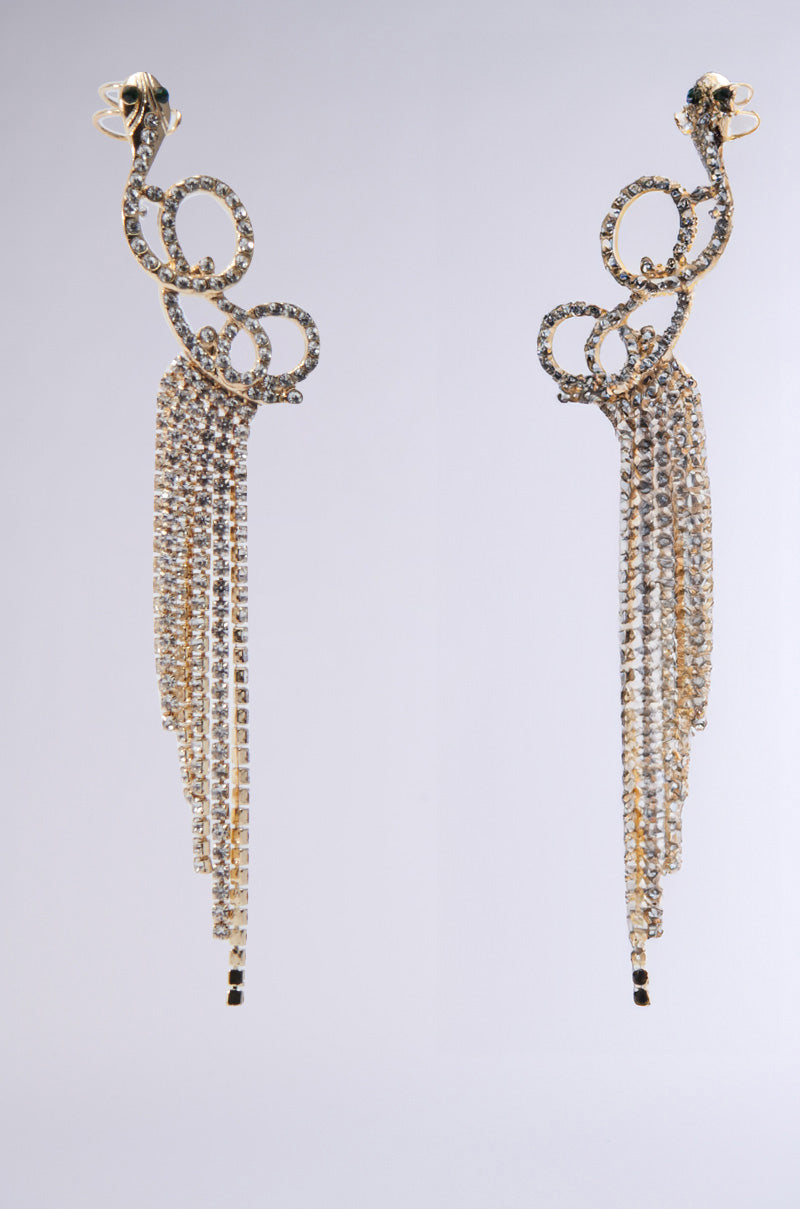 A pair of GALA GLAM EARRINGS embellished with swirling designs and cascading strands of sparkling crystals, featuring delicate snake silhouettes and rhinestone accents, set against a plain gray background.