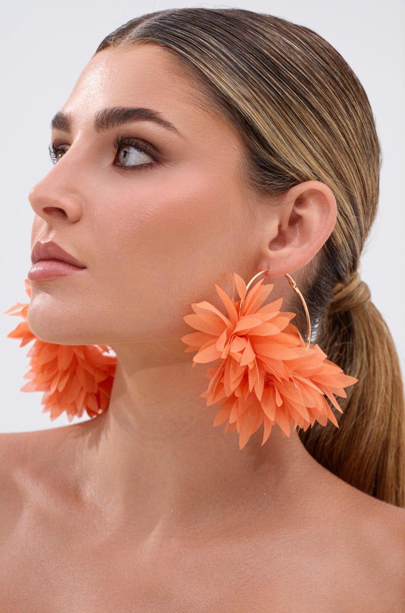 A woman with slicked-back brown hair wears the FOR THE SUMMER EARRING—oversized hoops featuring orange, flower-shaped embellishments. She gazes upwards against a simple, light background. 1