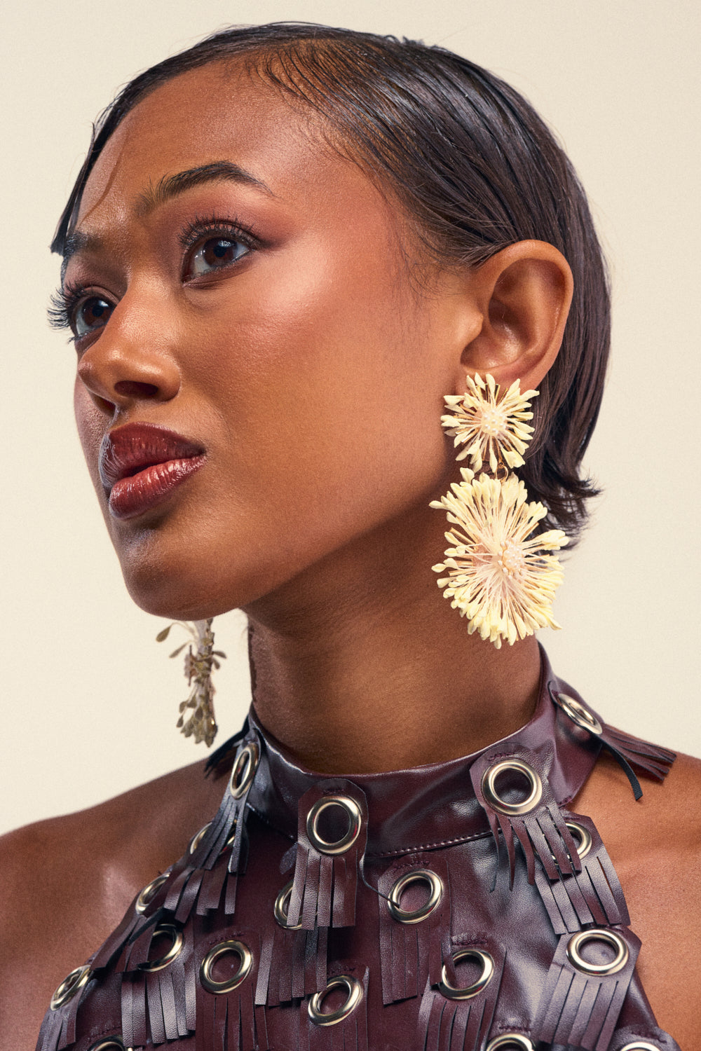A woman with short, sleek hair wears the ON HIS YACHT STATEMENT EARRING—large, cream-colored floral earrings—and a brown leather top with metal grommets, gazing upward confidently against a plain background.