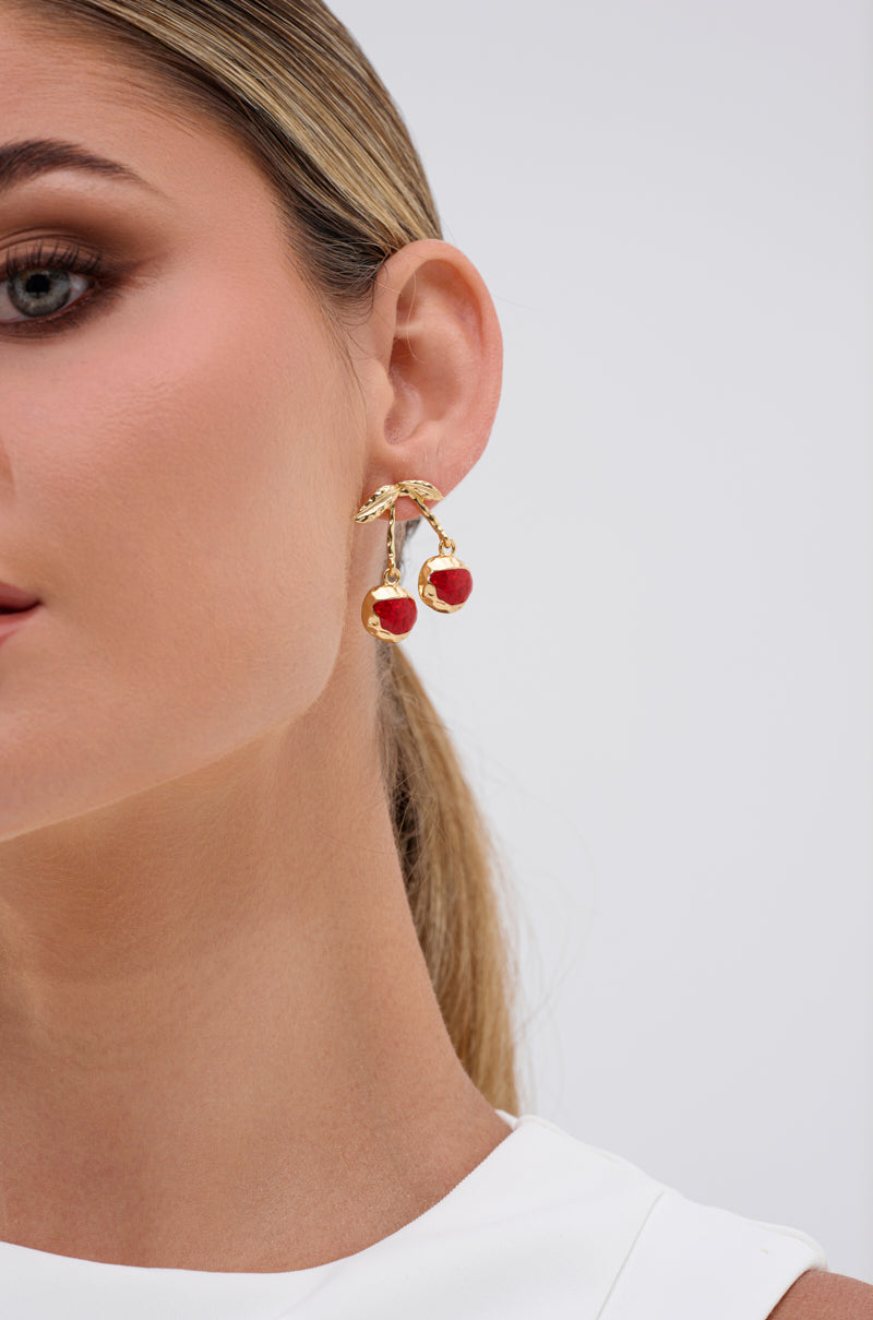 A close-up of a woman with light skin and straight blonde hair pulled back wearing POP MINE EARRING—jewel-embellished cherry earrings with gold-tone details and red enamel accents—styled with a sleeveless white top against a plain background. 1