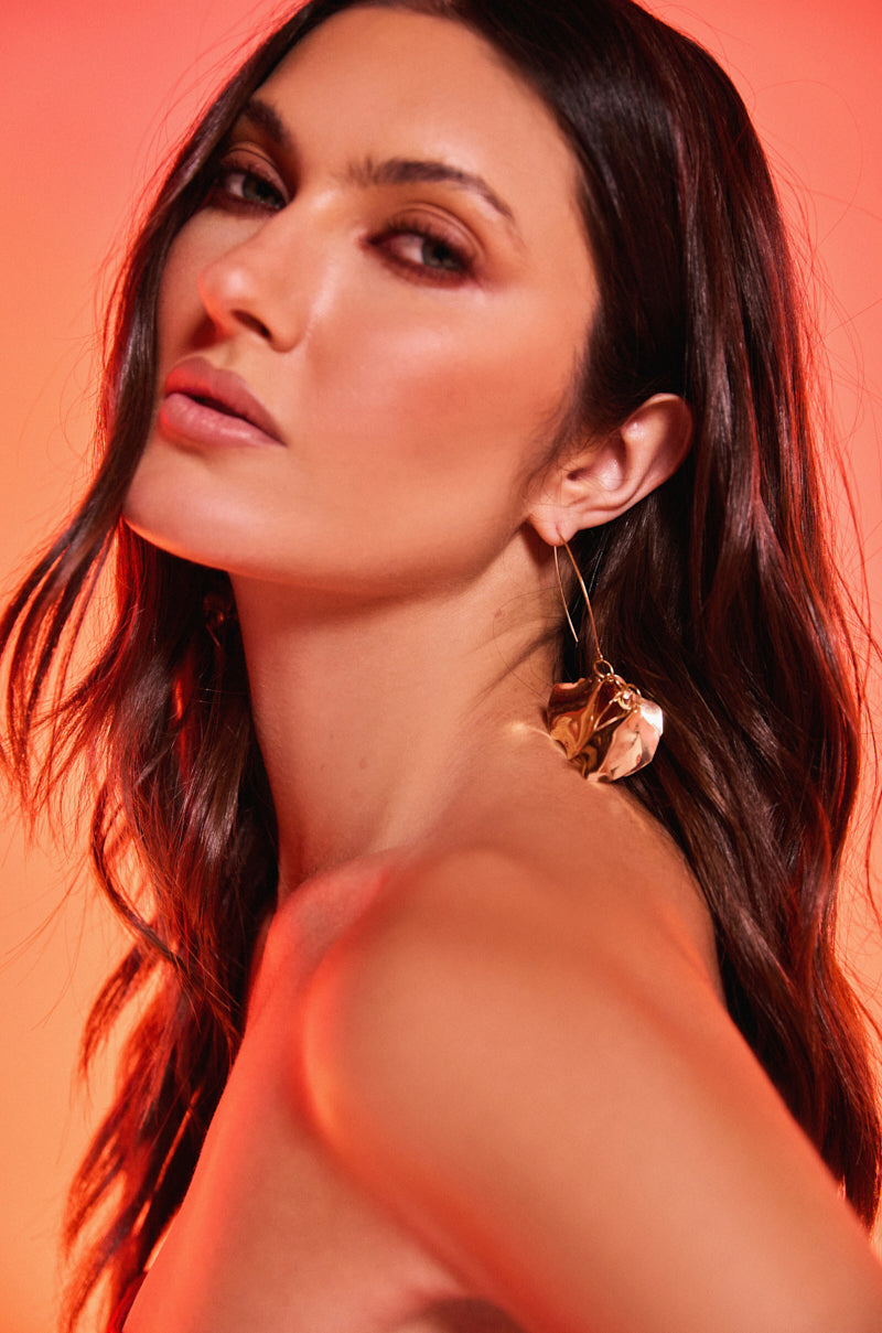 A woman with long, wavy brown hair poses against a red-orange backdrop, confidently wearing the METALLIC MANIA EARRING in gold and gazing at the camera with a soft, neutral expression. 1