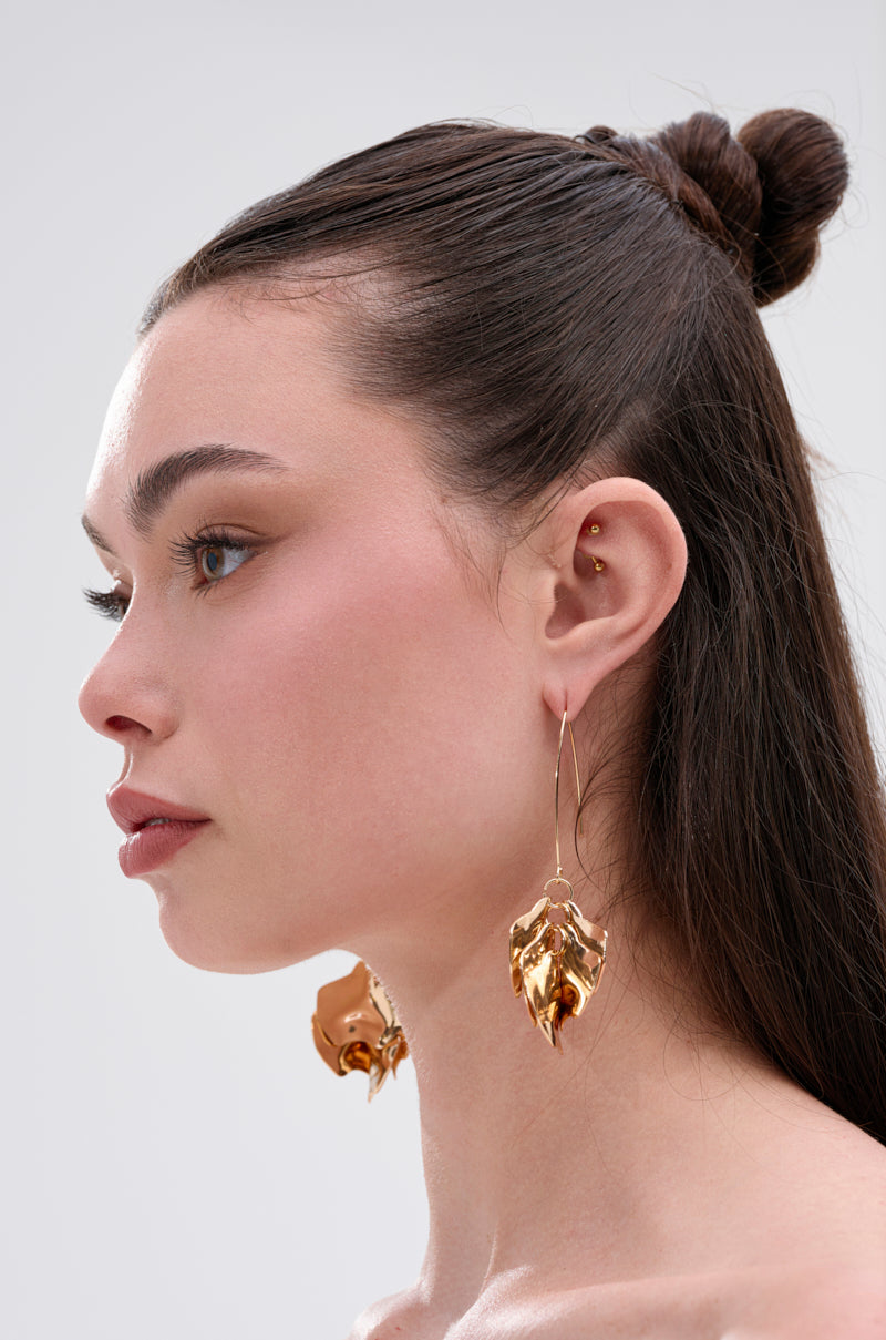 A woman with long brown hair in a bun wears large, crinkled METALLIC MANIA EARRINGS. Shown in profile against a plain light gray background, she has natural, minimal makeup. 3