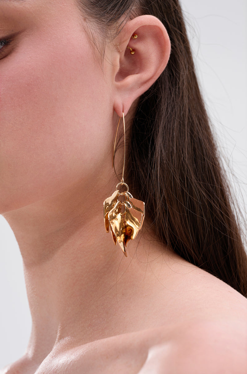A close-up of a woman's ear wearing the METALLIC MANIA EARRING, a large gold piece with a crinkled texture. She has multiple cartilage piercings, straight brown hair, and bare shoulders. 0