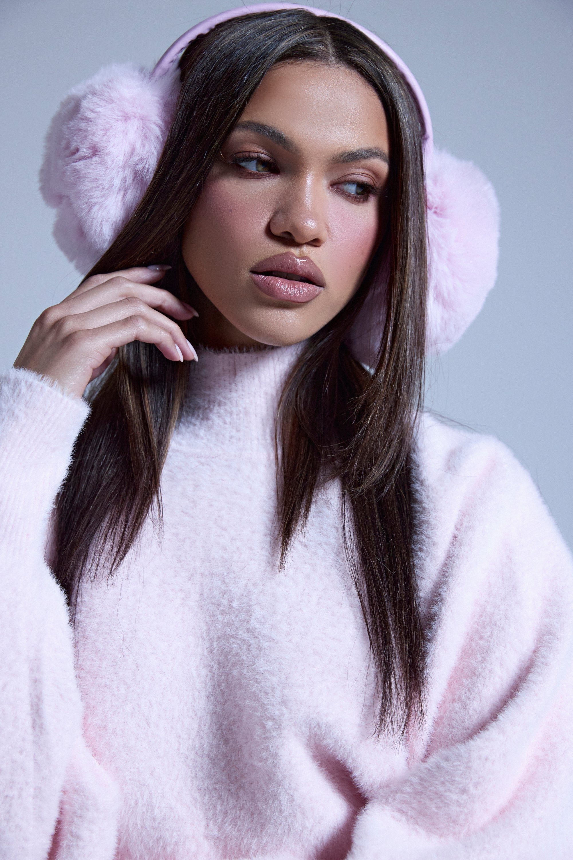 A woman with long brown hair and natural makeup gently touches her BB GURLLLL FAUX FUR EARMUFF in baby pink, paired with a matching sweater—an effortlessly chic winter accessory for style lovers. 0