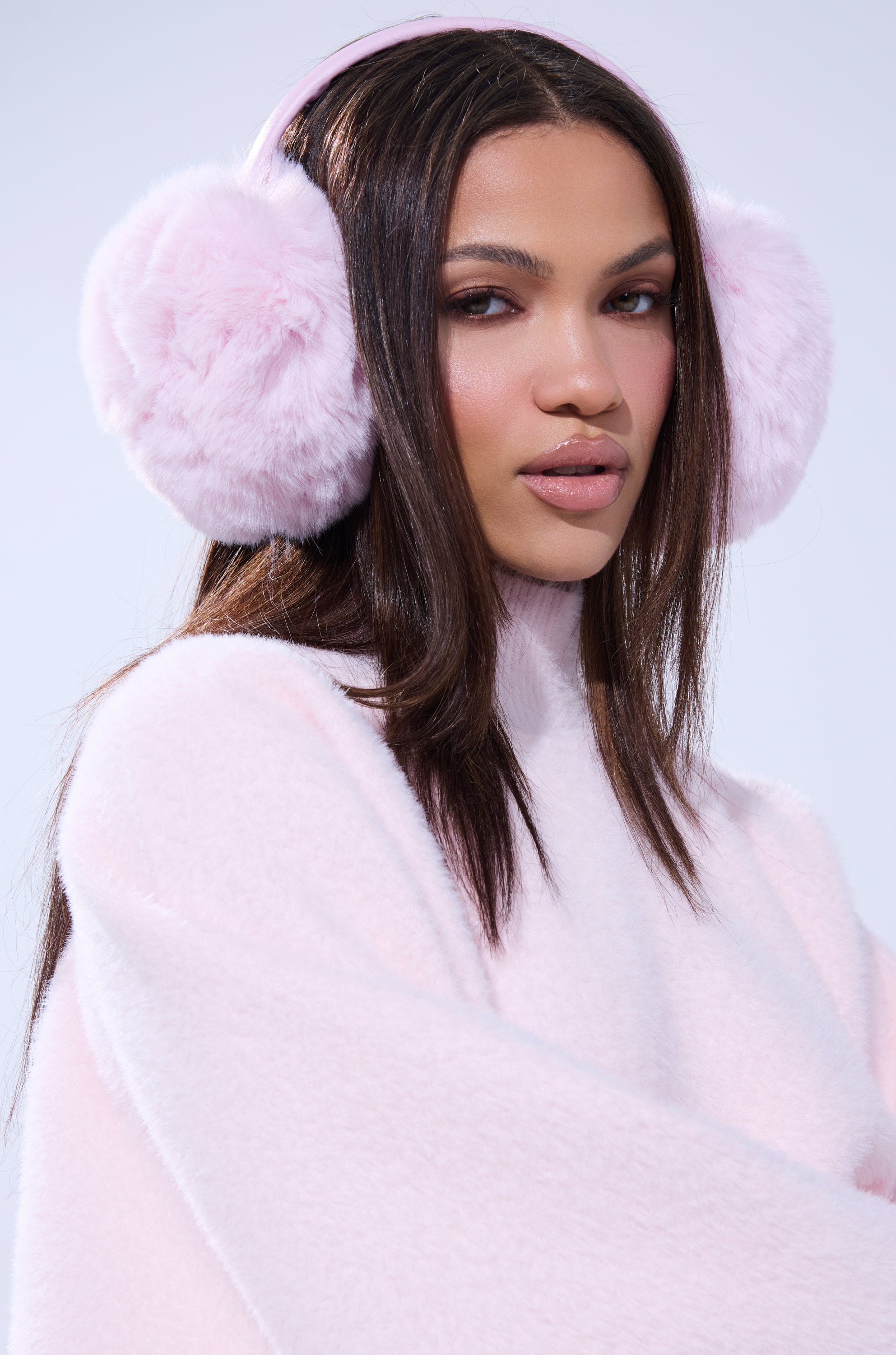 A woman with long brown hair wears BB GURLLLL FAUX FUR EARMUFF and a soft pale pink sweater, posing against a light background. 1