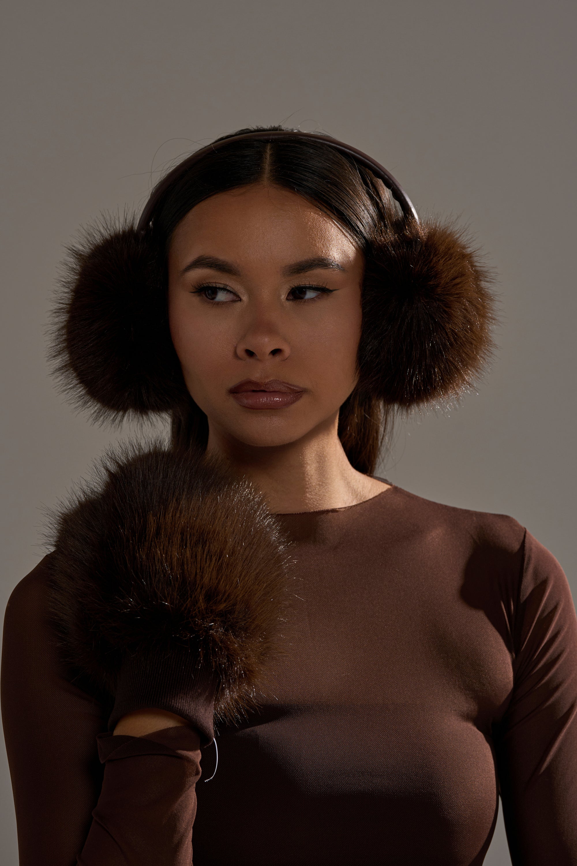 A woman models DARK CHOCOLATE FAUX FUR EARMUFFS with matching cuffs, paired with a long-sleeved brown top for a stylish, cozy look—perfect for fans of chic cold weather accessories. 2