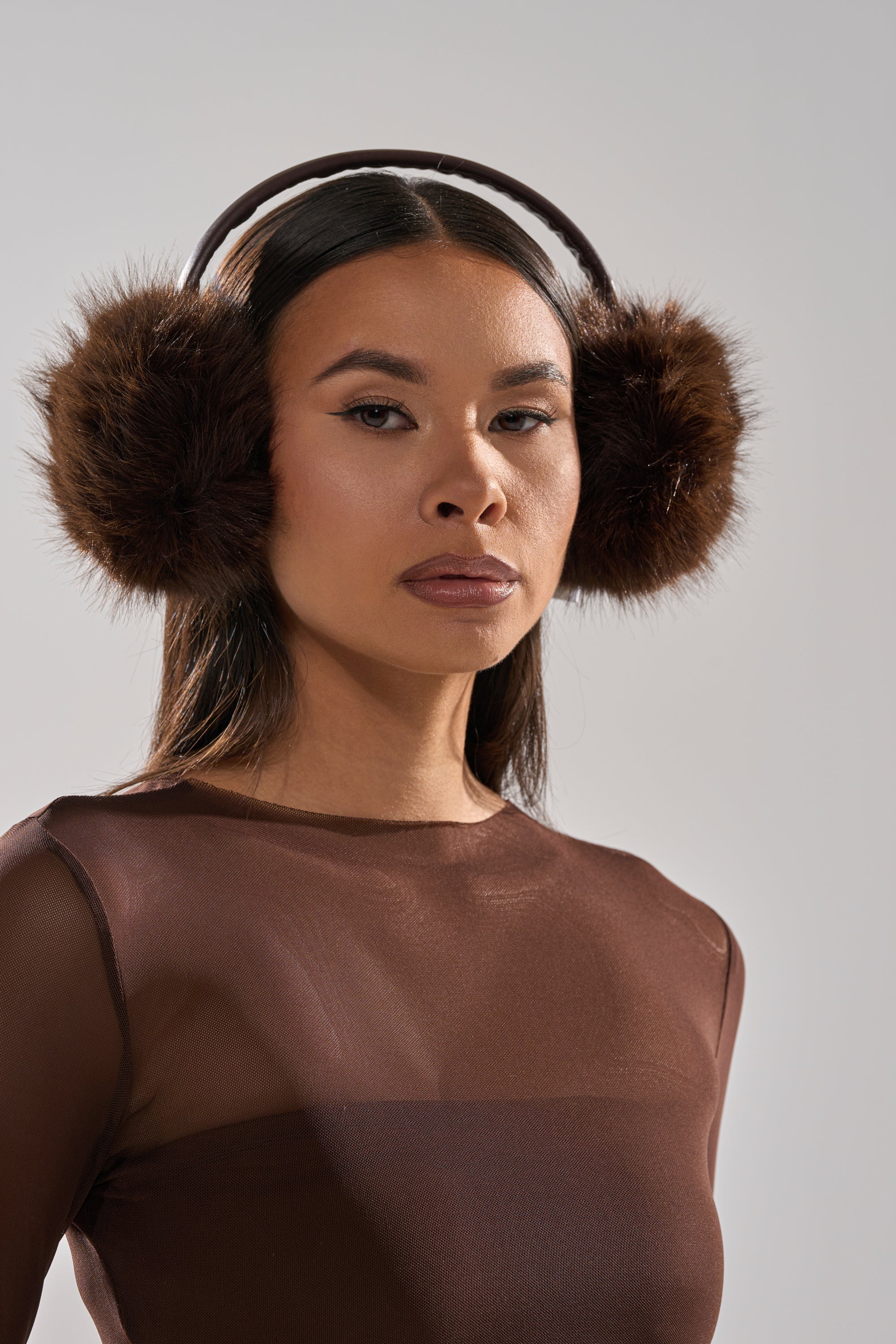 A woman models DARK CHOCOLATE FAUX FUR EARMUFFS and a sheer brown top, gazing at the camera against a light gray background for a cozy cold weather look. 1