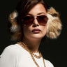 A person with short brown hair is seen wearing large WILD ONE FAUX FUR EAR MUFFS, aviator sunglasses, and a white sweater, complemented by a gold chain necklace. The dark background highlights the fashion-forward winter accessory.