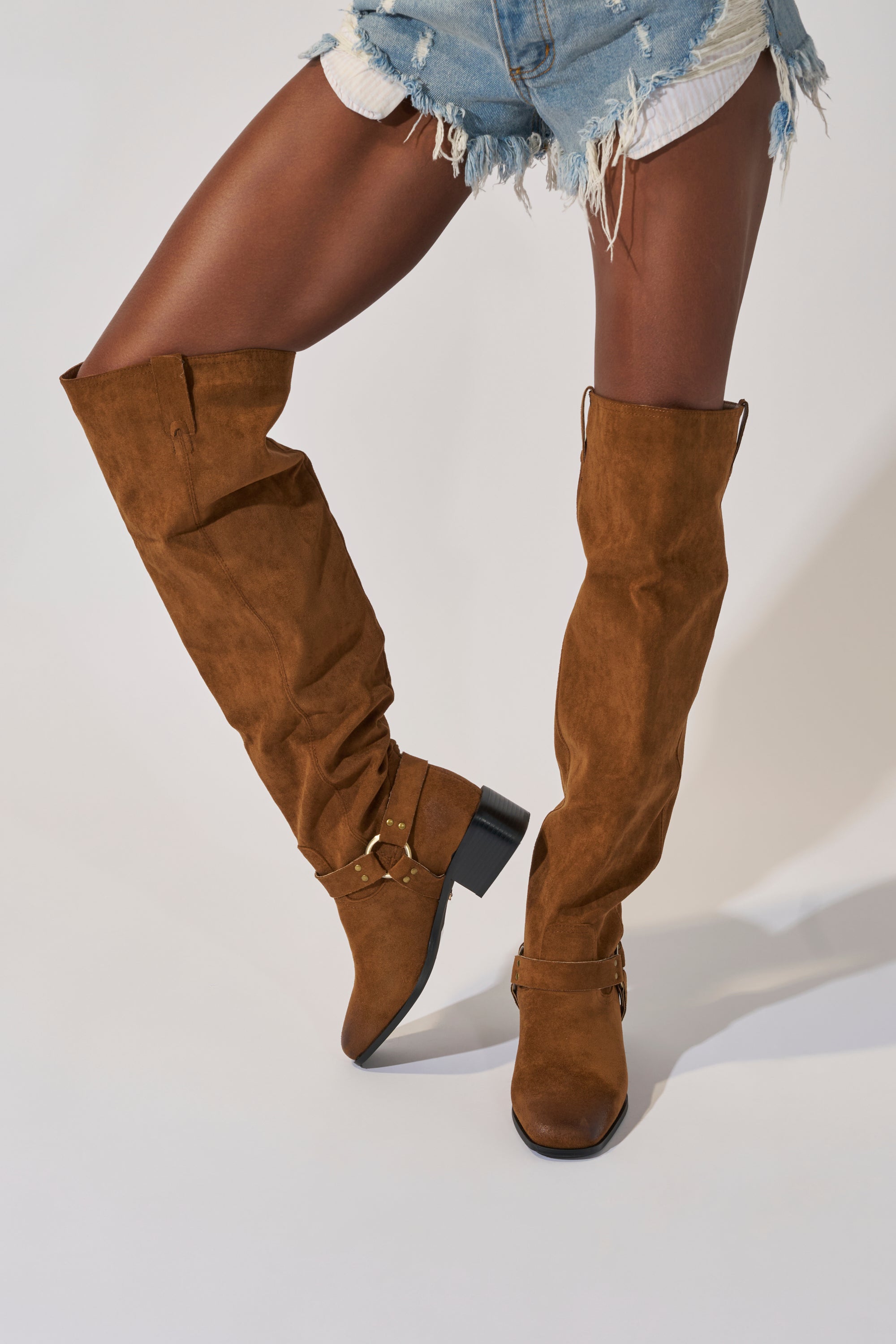 A person wearing frayed denim shorts and AZALEA WANG FLAT OVER THE KNEE RIDING BOOT in brown faux suede with black heels and gold buckle details, standing against a plain light background.