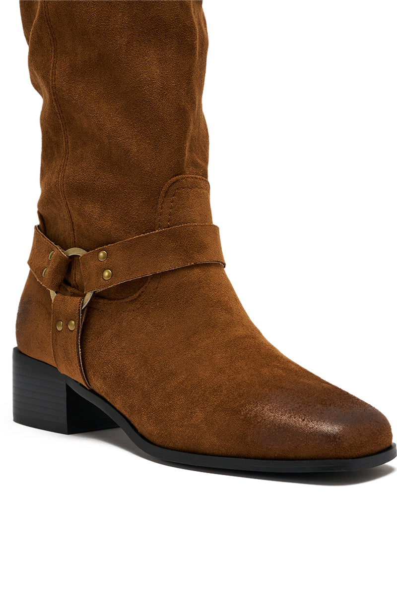 A close-up of the AZALEA WANG FLAT OVER THE KNEE RIDING BOOT in brown faux suede, featuring a low chunky heel, decorative straps, and brass studs, displayed on a white background.