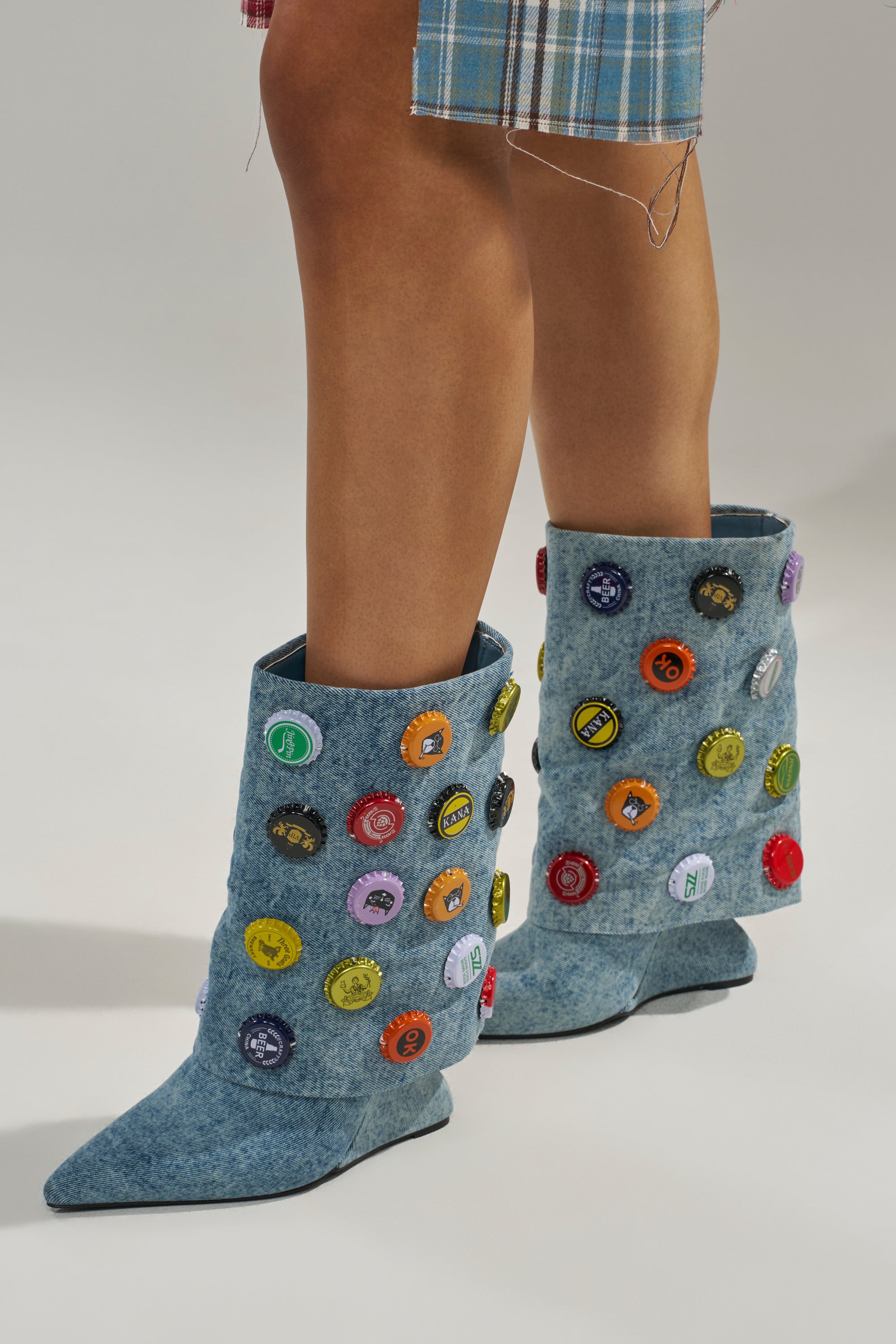 A person wears the AZALEA WANG EAVEN WEDGE EMBELLISHED DENIM BOOTIE—light blue booties with colorful patches and a slight wedge heel—paired stylishly with a blue plaid skirt featuring a trendy frayed hem.