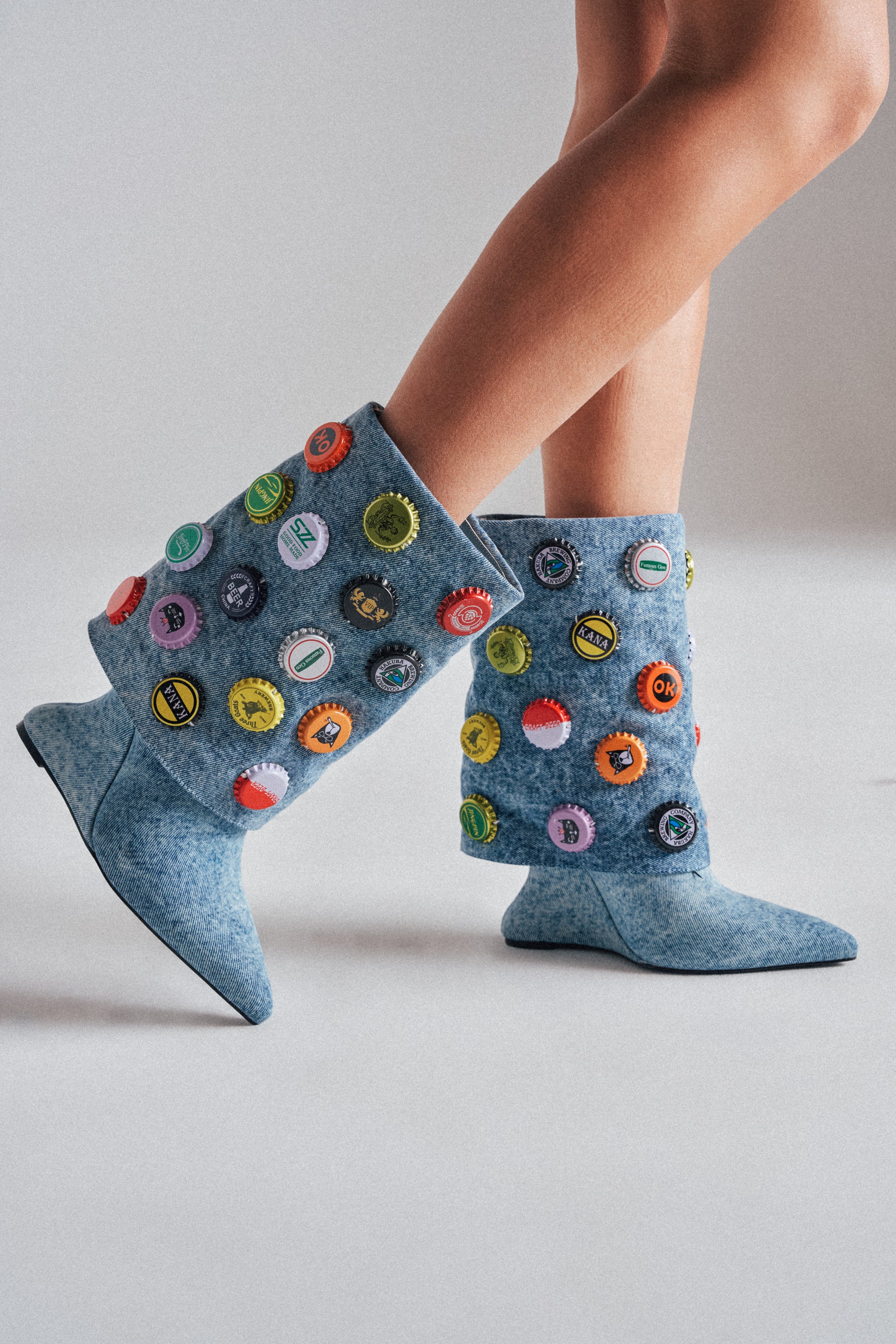 Someone stands on a light gray surface wearing only the AZALEA WANG EAVEN WEDGE EMBELLISHED DENIM BOOTIE—light blue denim booties with pointed toes and colorful bottle cap accents. Only their lower legs are visible. 0