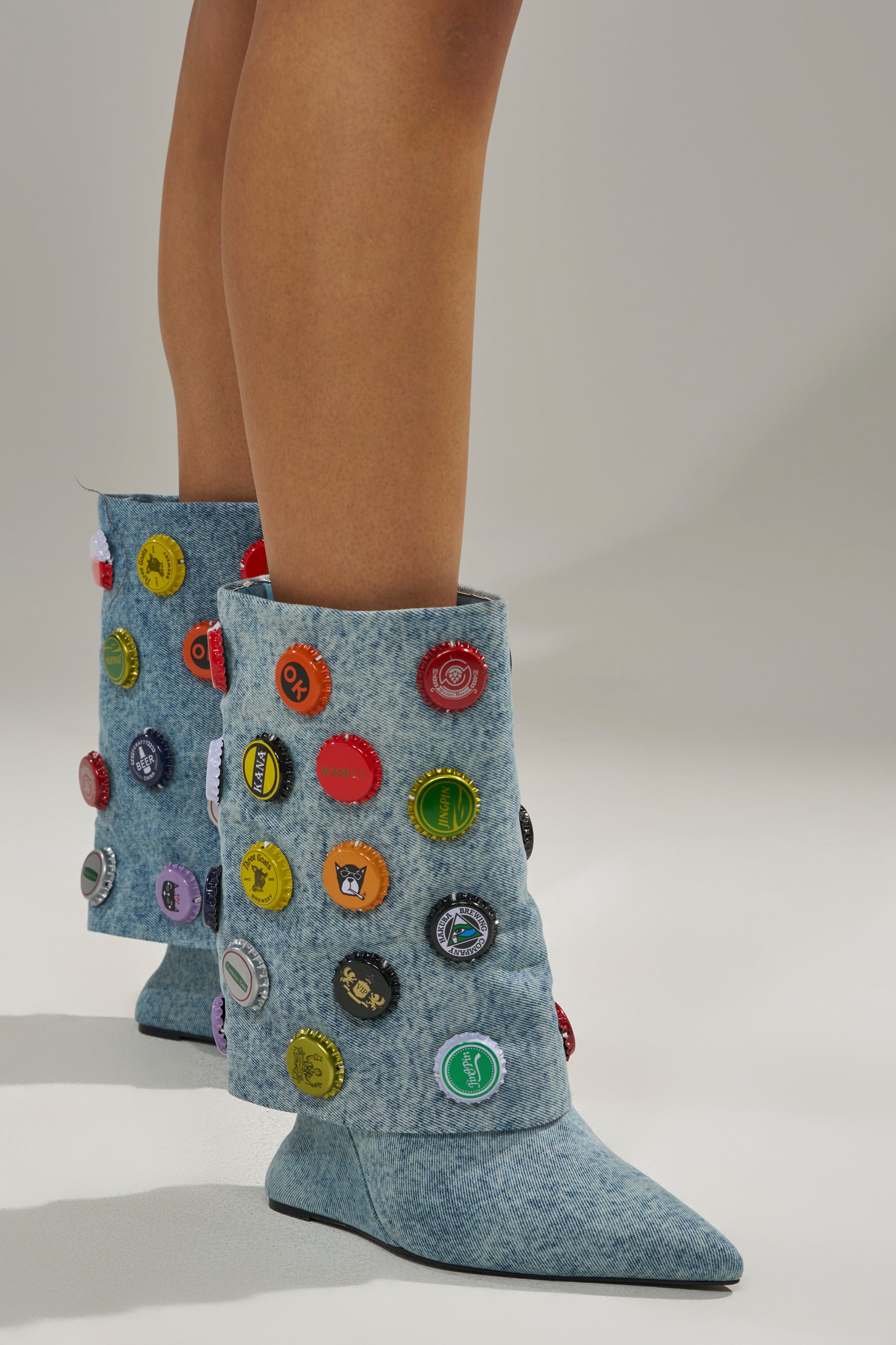 A model wears the AZALEA WANG EAVEN WEDGE EMBELLISHED DENIM BOOTIE—light blue, textured denim booties with wedge heels, colorful bottle caps, and patches, reaching mid-calf and shown against a neutral background.