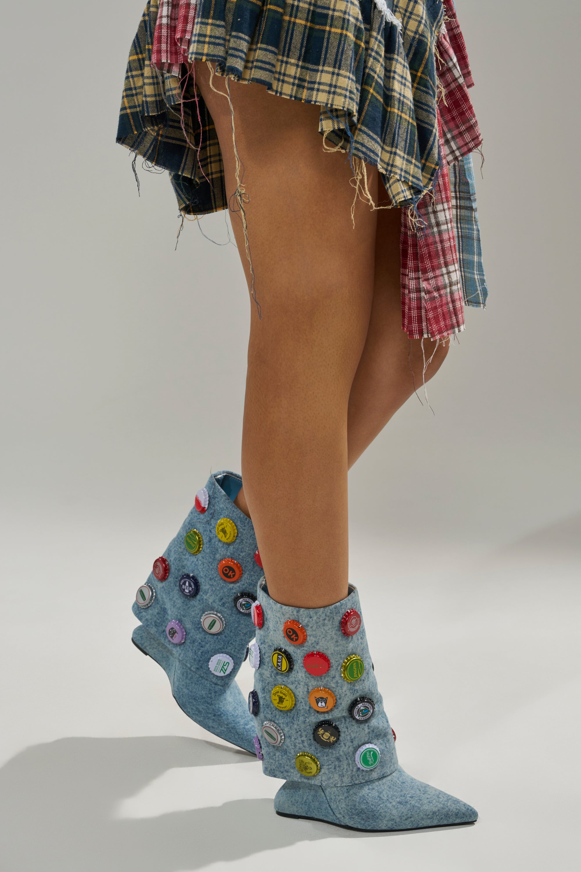 A person stands against a plain background, wearing a patchwork plaid skirt and AZALEA WANG EAVEN WEDGE EMBELLISHED DENIM BOOTIE adorned with colorful bottle caps.