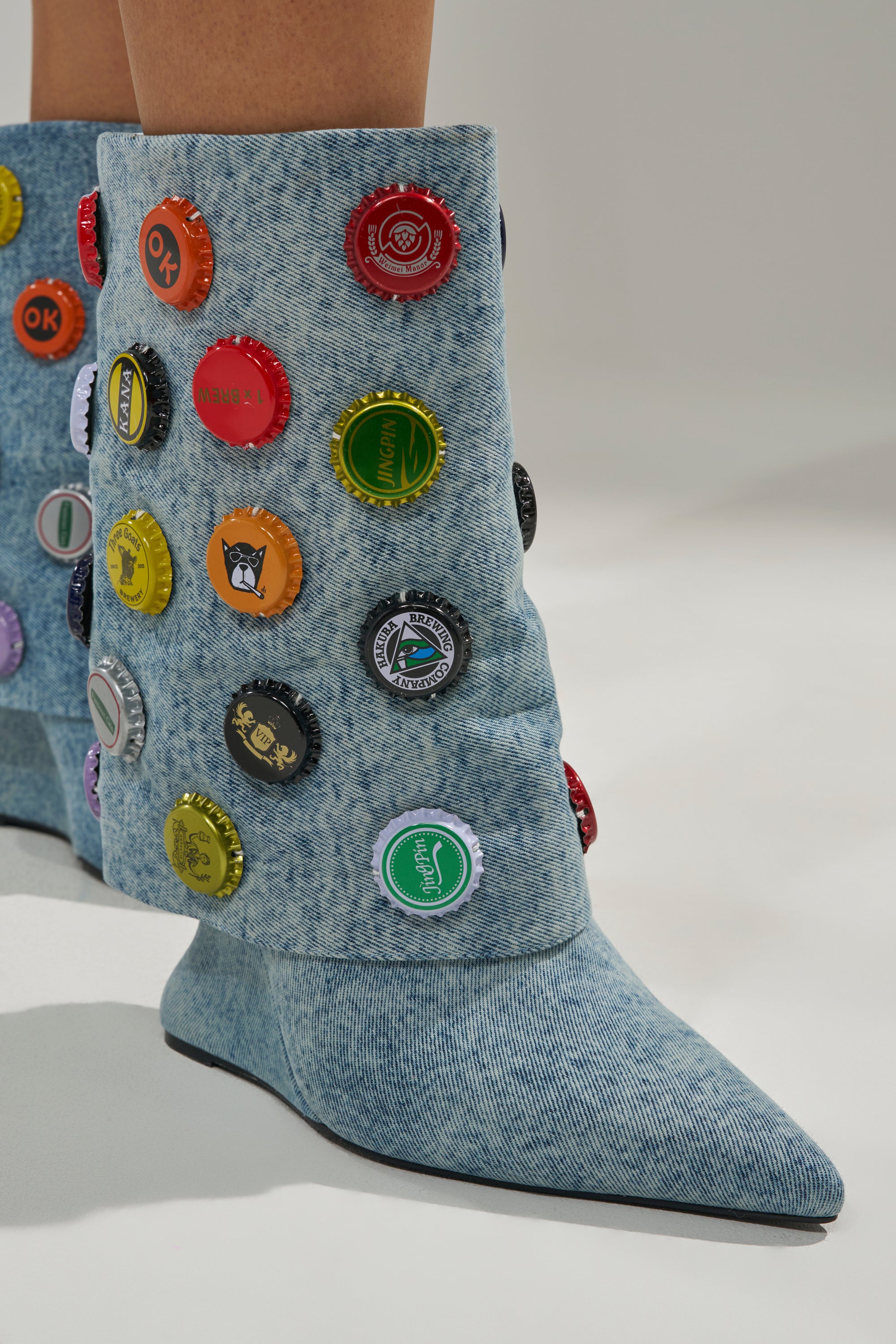 The AZALEA WANG EAVEN WEDGE EMBELLISHED DENIM BOOTIE features light blue denim and a wedge heel, fully covered in rows of colorful bottle caps, shown worn on a white surface.