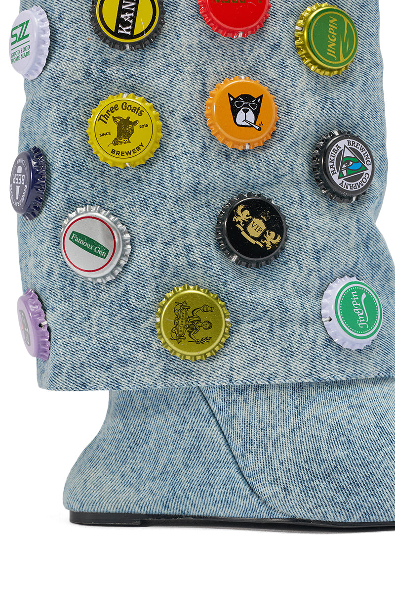 The AZALEA WANG EAVEN WEDGE EMBELLISHED DENIM BOOTIE features a close-up of denim adorned with colorful bottle caps showcasing various logos and designs, completed with a chunky wedge heel against a clean white background.