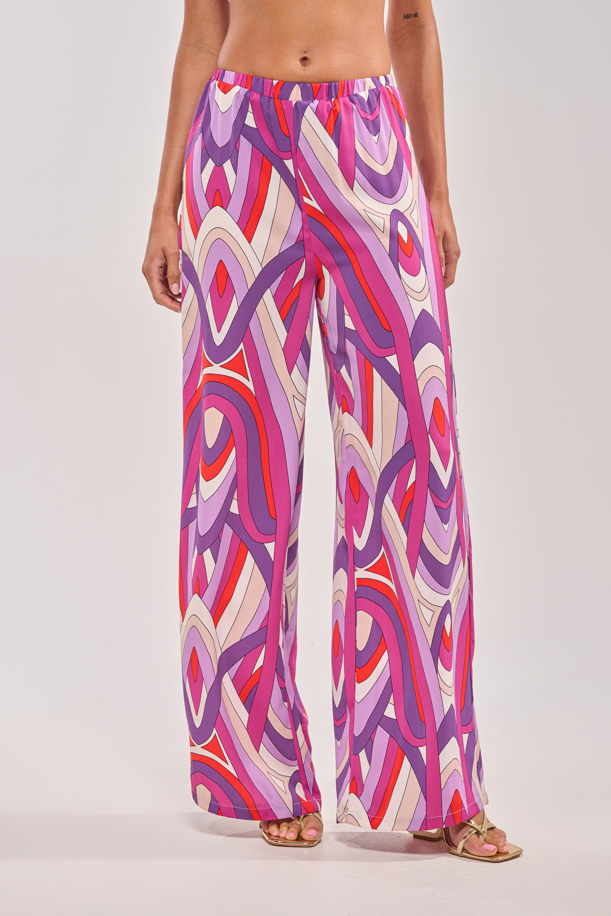 A person stands against a plain background wearing the GIRLY POP PALAZZO PANT, featuring a purple geometric print in shades of pink, purple, red, and white, with a high-rise wide-leg fit. 0