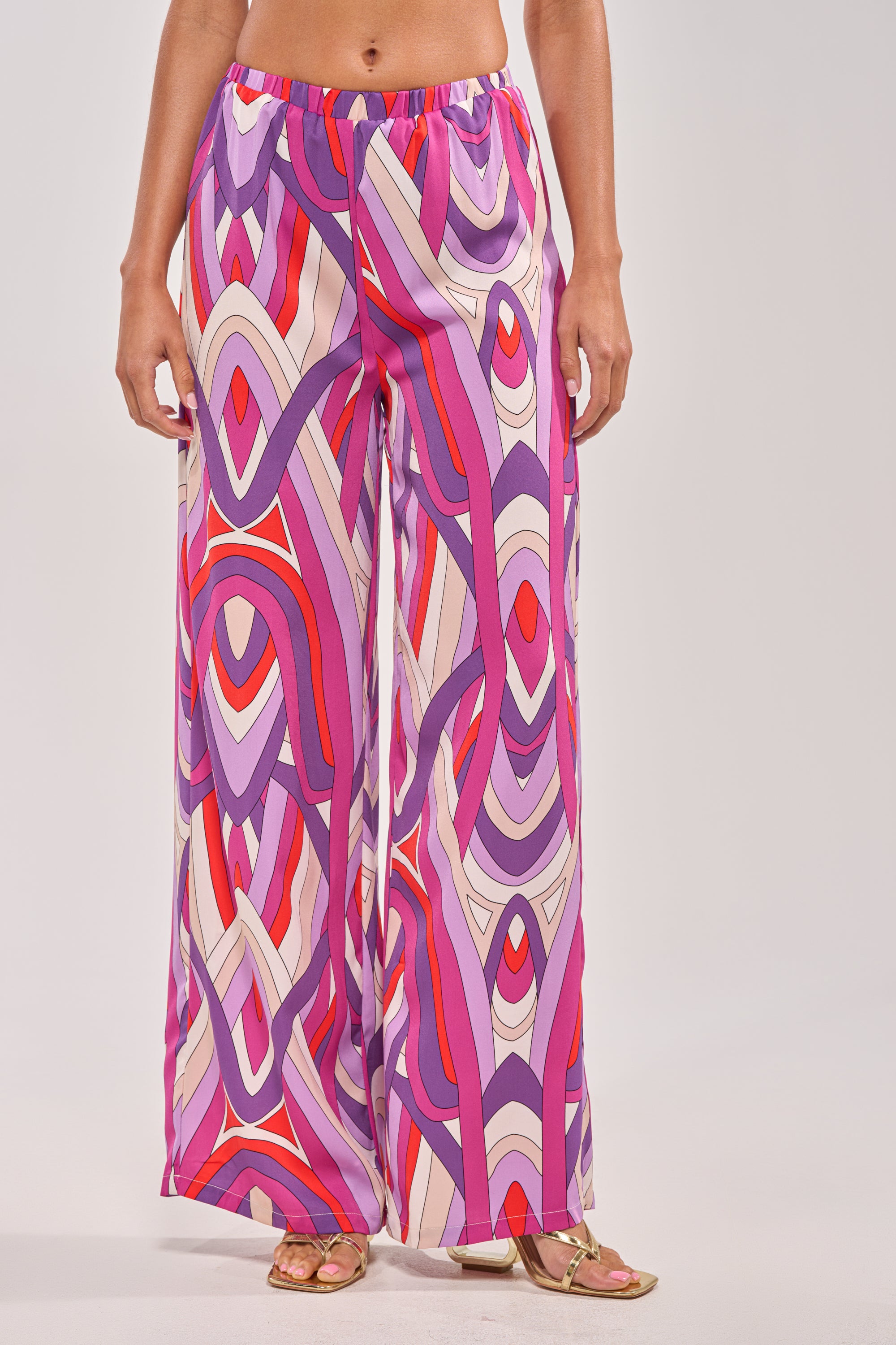Someone stands against a plain background wearing the GIRLY POP PALAZZO PANT—high-rise, wide-leg pants with a vibrant geometric pattern in purple, pink, red, and white—paired with gold sandals. 1