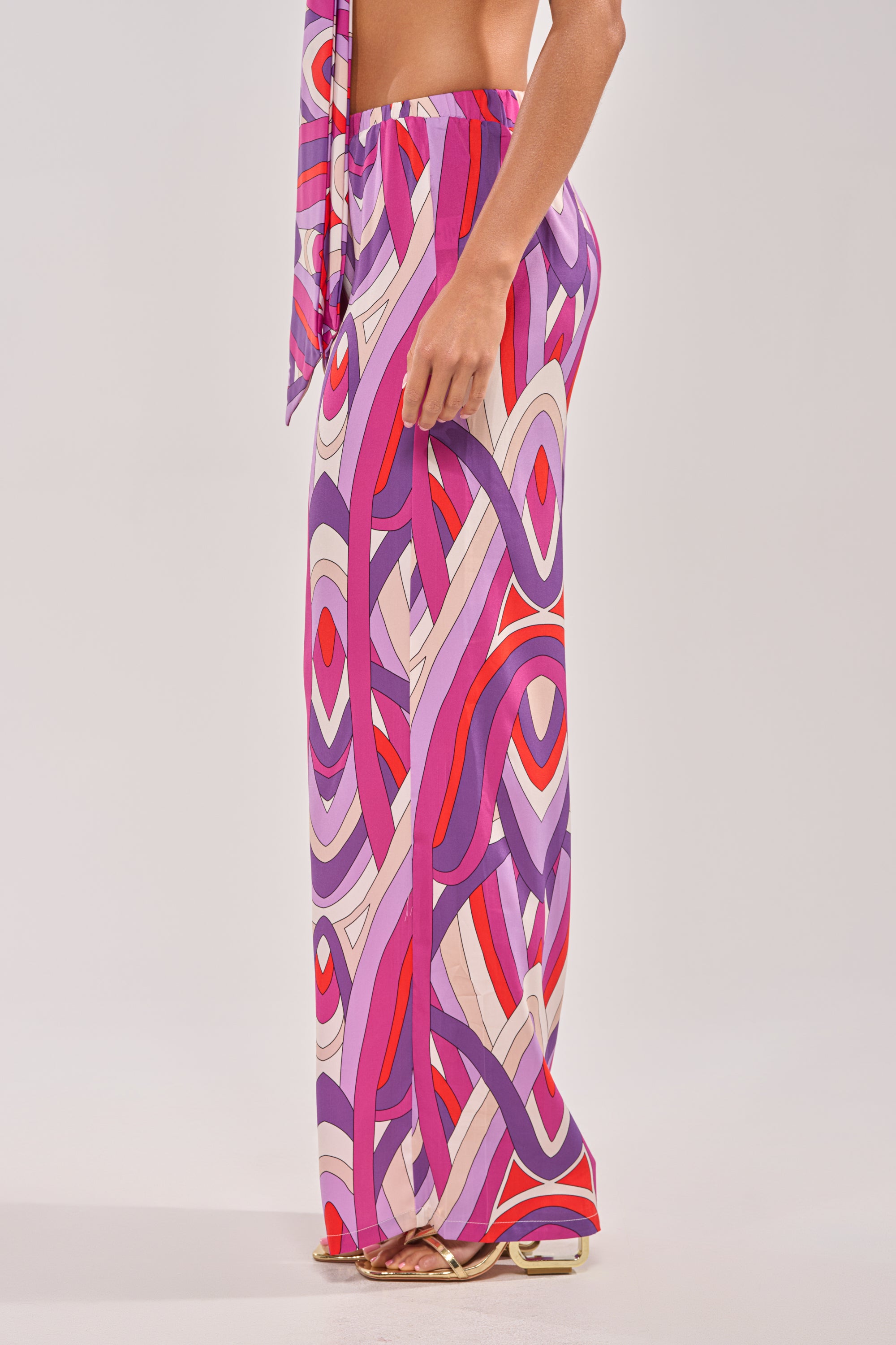 Someone stands sideways against a plain background wearing the GIRLY POP PALAZZO PANT, featuring a purple base with a colorful abstract geometric pattern in shades of pink, red, and white. 2
