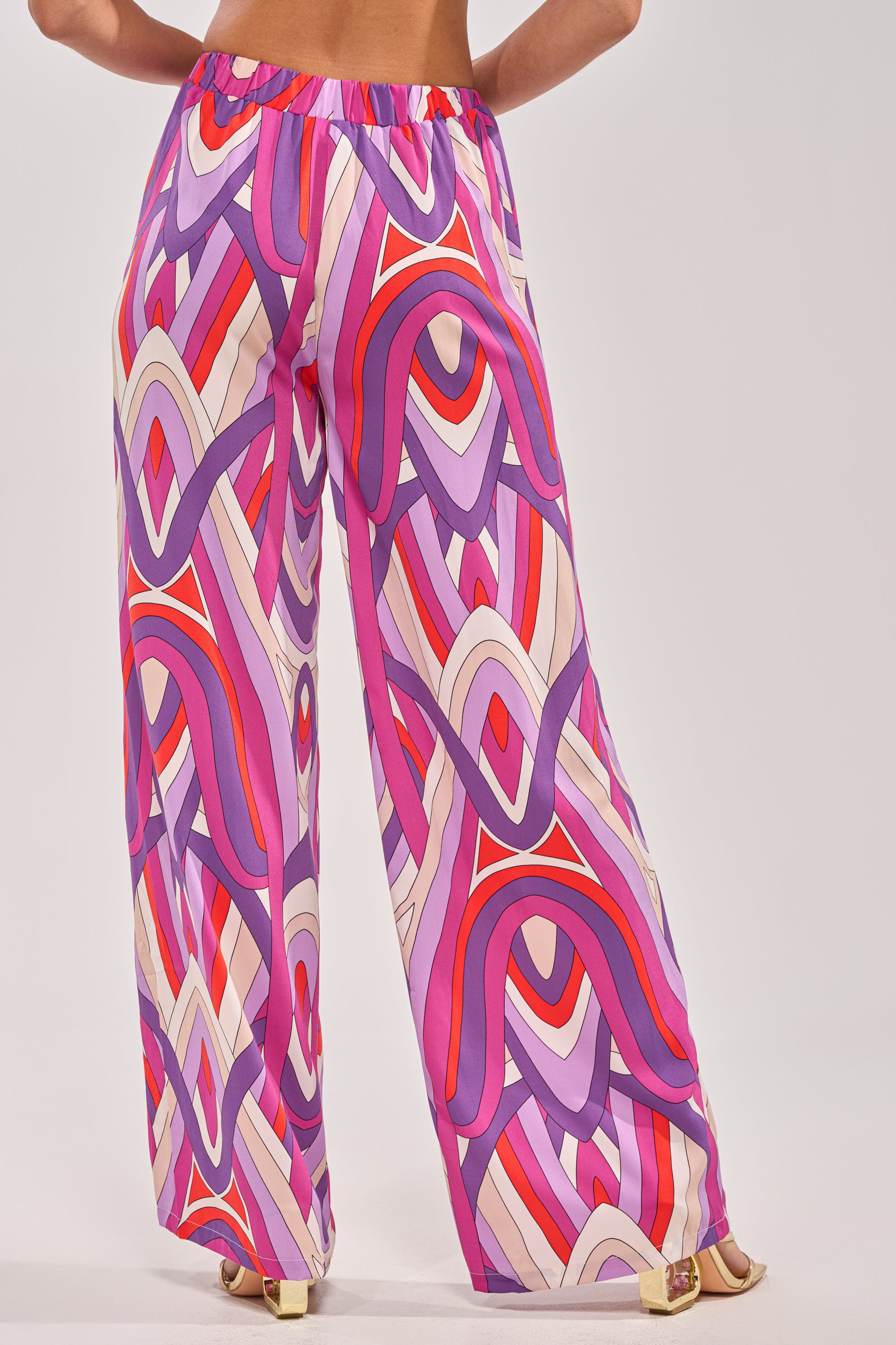A person is seen from the back wearing GIRLY POP PALAZZO PANT featuring a bold geometric pattern in purple, red, pink, and cream, paired with gold open-toe heels. 3