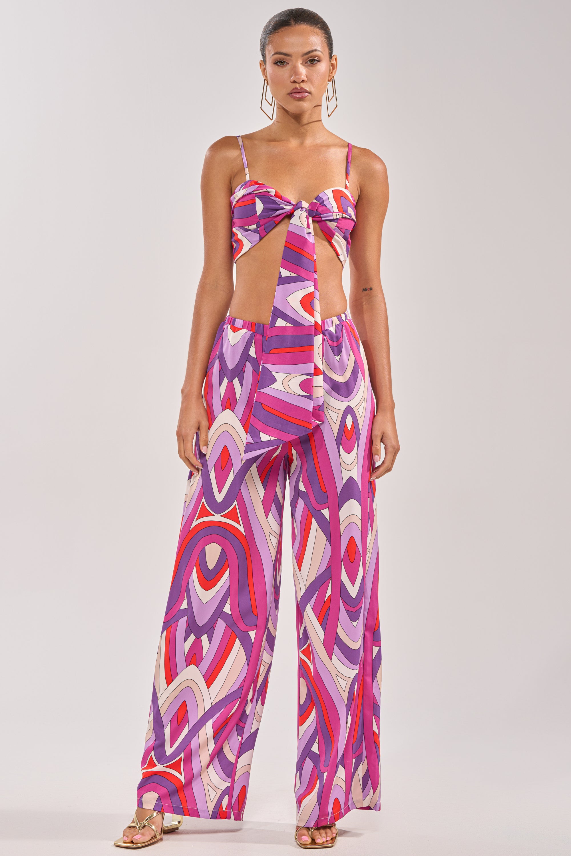 A woman wears the GIRLY POP PALAZZO PANT, featuring a purple geometric print in pink, purple, red, and white, styled with a matching sleeveless crop top, sandals, and geometric earrings. 4