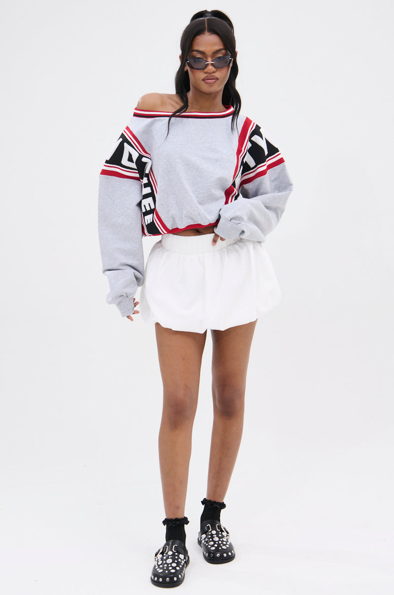 A person wears an off-shoulder gray sweatshirt with red and black accents, a BUBBLE BUBBLE BUBBLE SKIRT in white with a shirred waistband, embellished black shoes, long hair, sunglasses, and black socks against a plain white background.