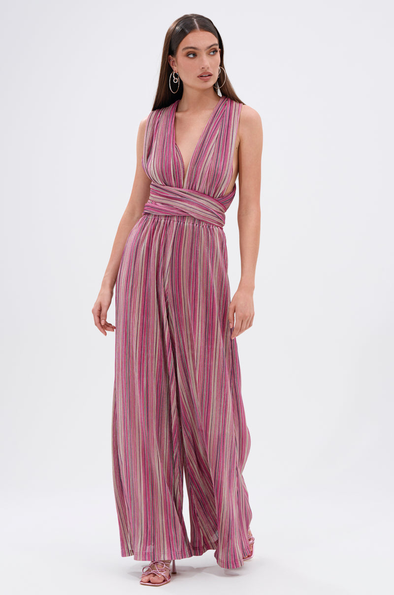 A woman models the JUMPIN AROUND SLINKY JUMPSUIT—a sleeveless, wide-leg piece with a deep V-neckline and waistband, featuring pink, purple, and white vertical stripes—showcased against a plain white background. 0