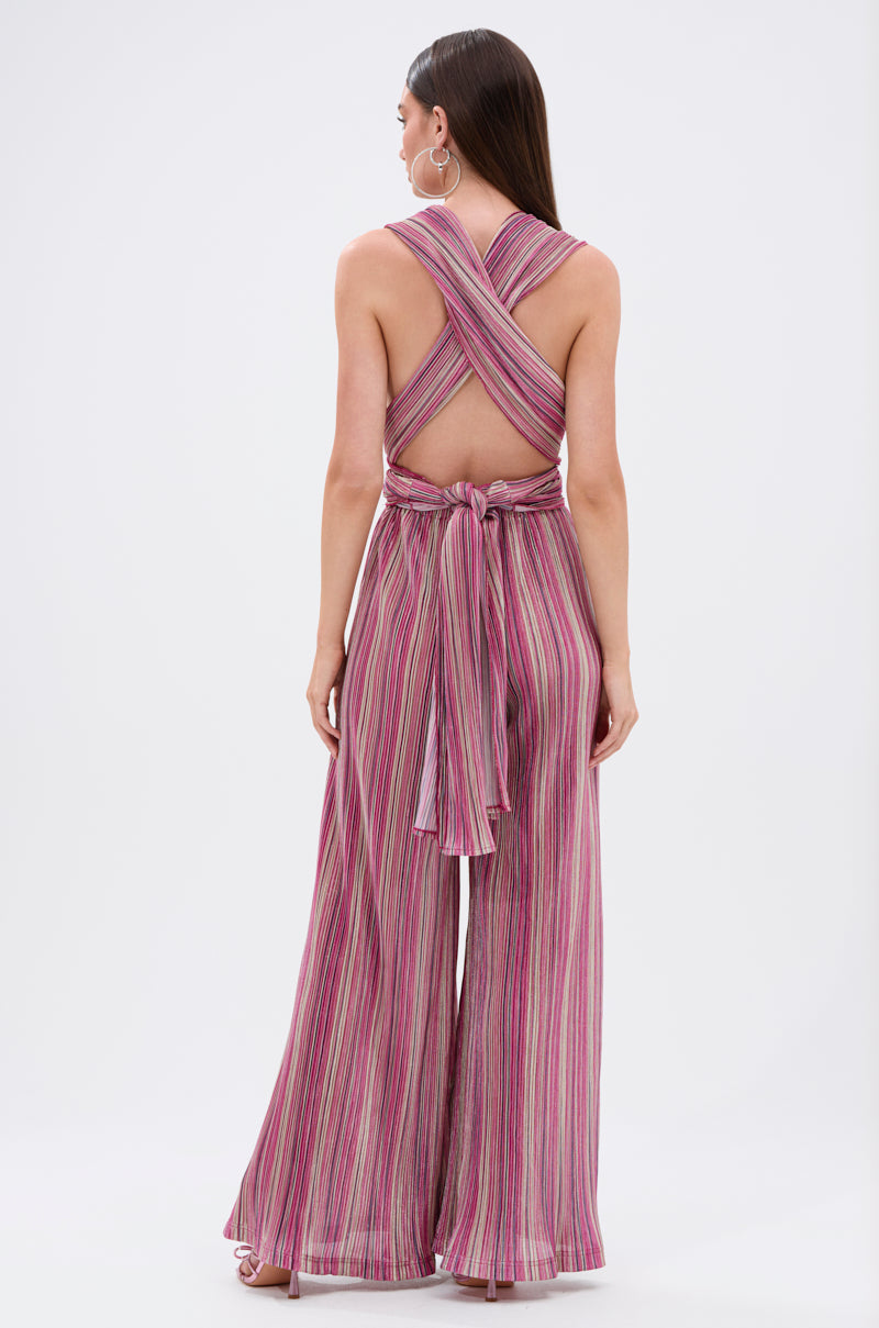 Woman stands with her back to the camera in the JUMPIN AROUND SLINKY JUMPSUIT—a sleeveless, convertible jumpsuit with wide-leg pants, pink, purple, and white stripes, crisscross straps, and a bow accent at the back waist. 9