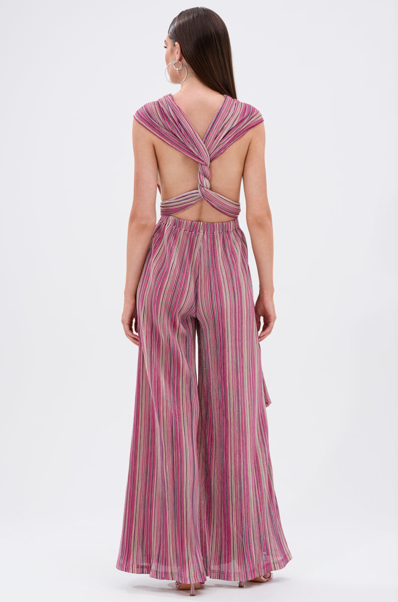 A woman faces away in the JUMPIN AROUND SLINKY JUMPSUIT, featuring pink, purple, and white vertical stripes, wide-leg pants for elegant flow, and a twisted knot accentuating the open back for a modern style. 10