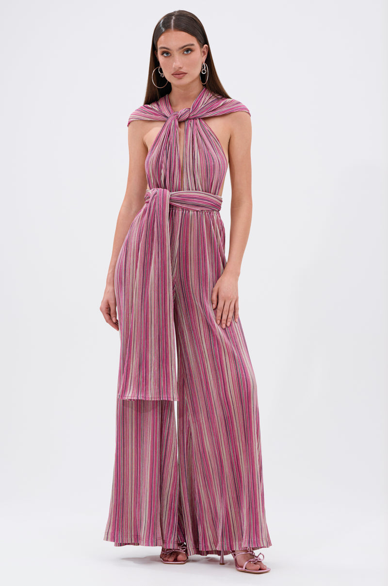 A woman wears the JUMPIN AROUND SLINKY JUMPSUIT, a sleeveless halter-neck jumpsuit with wide-leg pants, multicolored vertical stripes in pink and purple, and a matching waist tie. She has long straight hair and hoop earrings. 2