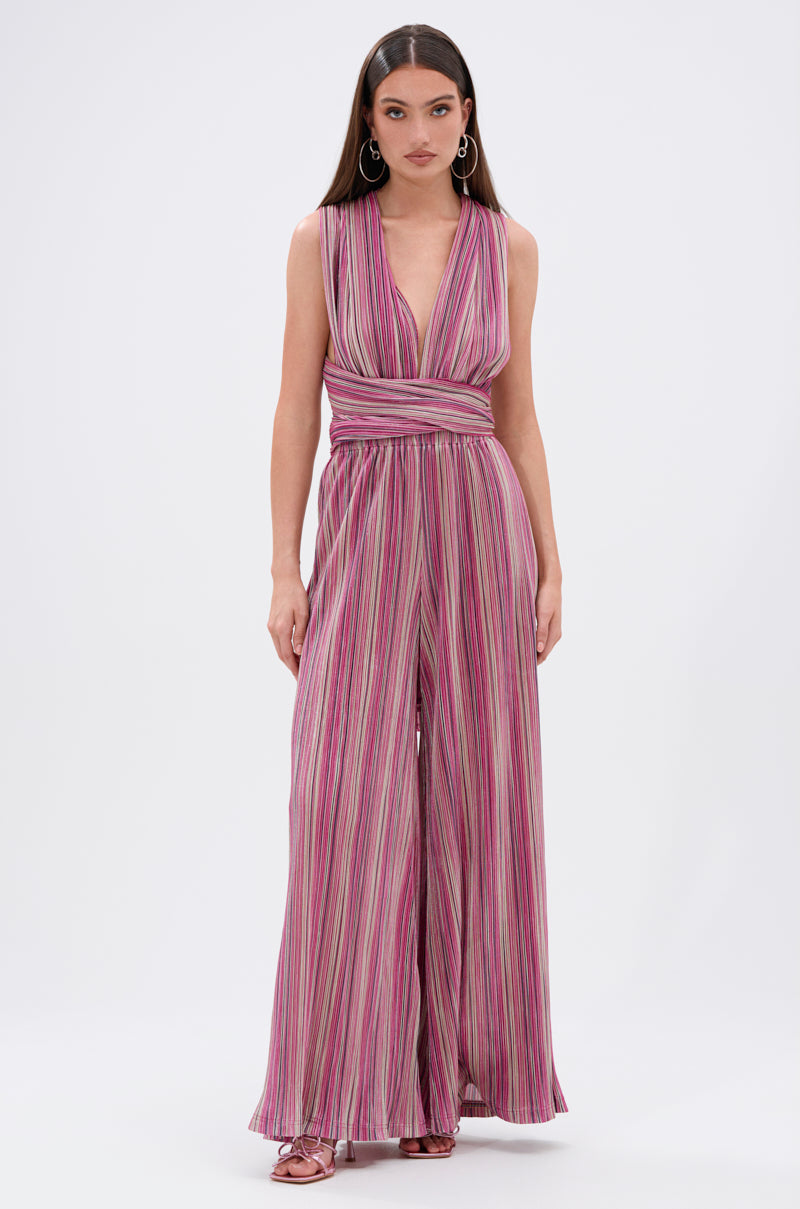 A woman wears the JUMPIN AROUND SLINKY JUMPSUIT, a sleeveless convertible style with wide-leg pants, vertical pink, purple, and white stripes, a deep V-neckline, and a cinched waist. She pairs it with hoop earrings and heeled sandals. 3