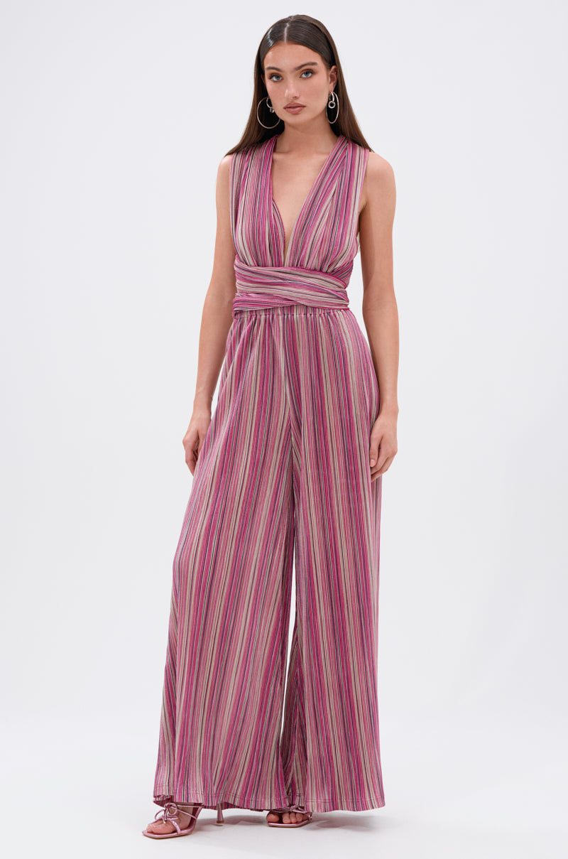 A woman stands against a white background wearing the JUMPIN AROUND SLINKY JUMPSUIT, a sleeveless convertible jumpsuit with a deep V-neck, pink and purple stripes, wide-leg pants, and a wrapped waist. 4