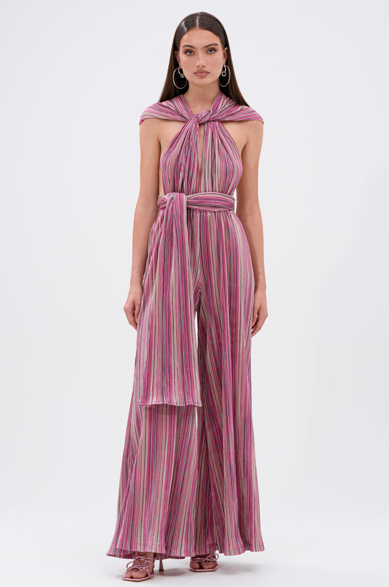 A woman models the JUMPIN AROUND SLINKY JUMPSUIT—sleeveless, convertible, with vertical pink, purple, and white stripes, a tied halter neckline, matching waist sash, and wide-leg pants—paired with heeled sandals against a white background. 6