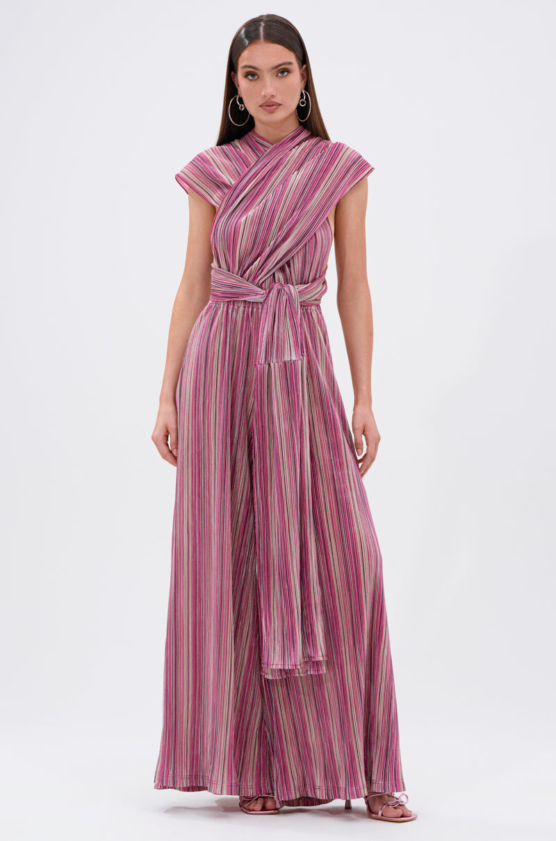 A woman with long brown hair wears the JUMPIN AROUND SLINKY JUMPSUIT, featuring pink, purple, and white stripes, wide legs, a crisscross wrap top, and a tied waist sash. She accessorizes with hoop earrings and sandals. 7