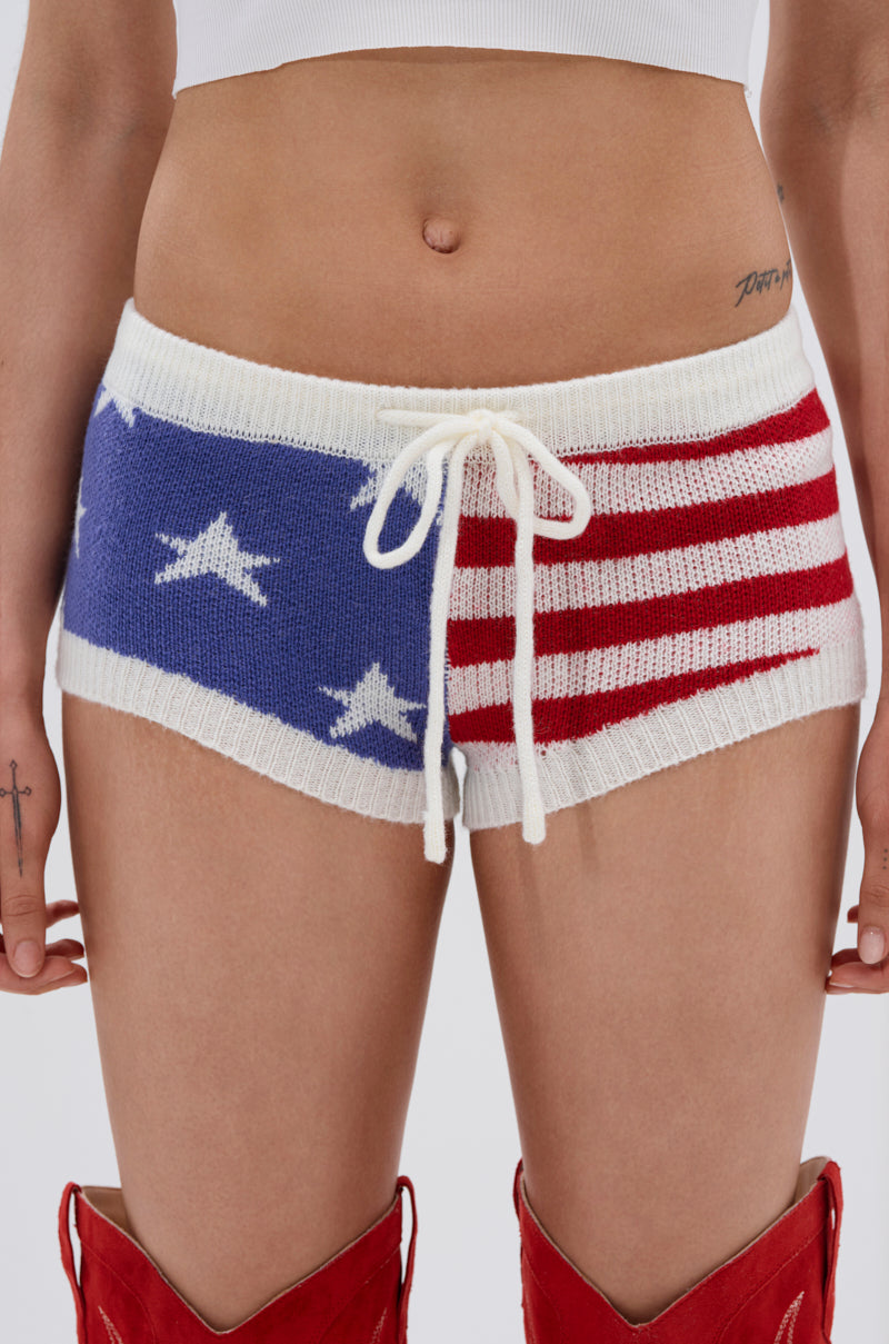 A person wearing the PARTY IN THE USA SHORT—knit shorts with an American flag pattern—shows off a "Dior" tattoo and bold red boots for a true USA-themed look.