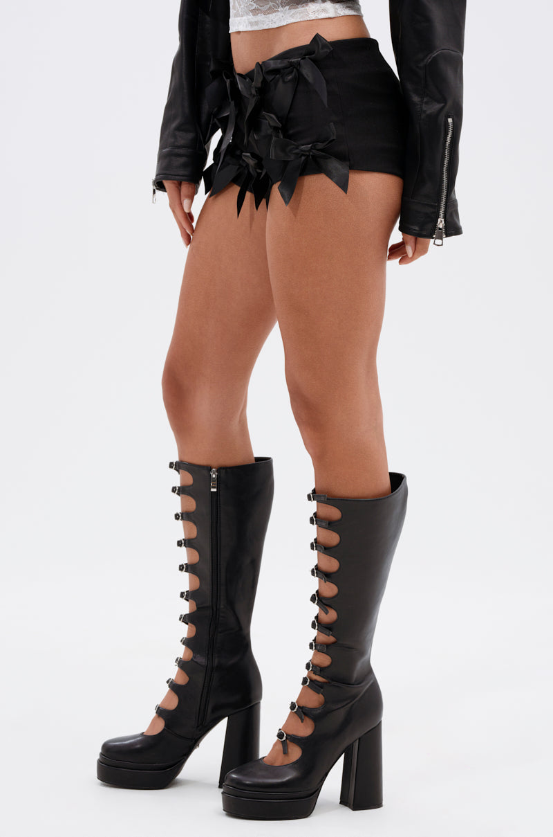 A person wearing a black leather jacket, CUTIE PIE SATIN BOW HOT SHORT with a curve-hugging fit, and black platform knee-high boots with button details stands on a plain white background. 4
