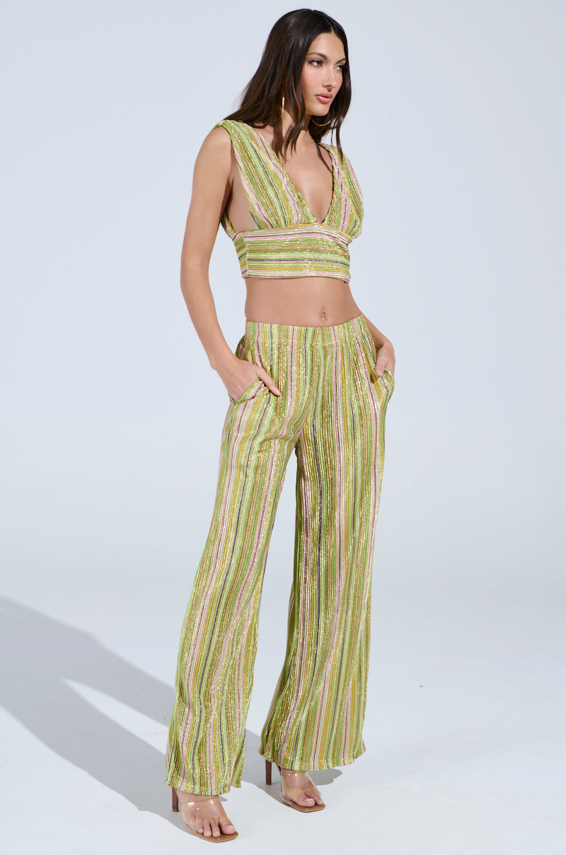 A confident woman, with long hair, poses against a light background wearing a sleeveless green top featuring a deep V-neck and multicolored stripes. She pairs it with high-rise "MIXED EMOTIONS PALAZZO PANT" and poses with her hands in her pockets for a vibrant ensemble.