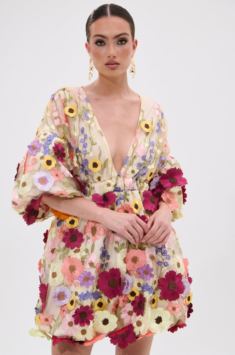 A woman models the FLORAL BABY MINI DRESS, featuring sheer fabric, colorful embroidered floral appliques, and a deep V-neckline. She accessorizes with gold hoop earrings and has her hair pulled back, standing against a white background. 1