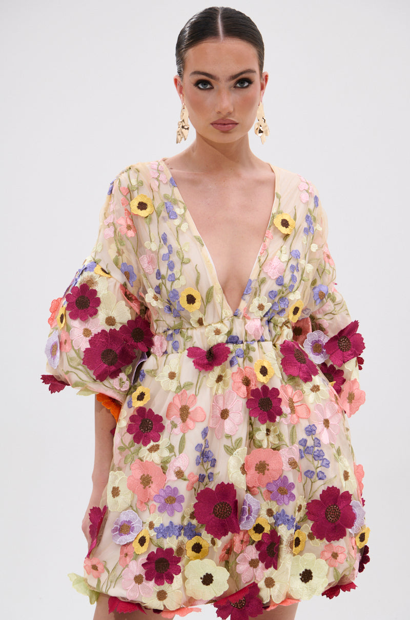 A woman with slicked-back hair and gold earrings models the FLORAL BABY MINI DRESS, featuring a deep V-neck, voluminous sleeves, and colorful embroidered flower appliques in pink, purple, yellow, and red against a plain background. 2