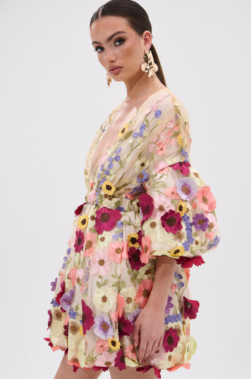 A woman showcases the FLORAL BABY MINI DRESS, styled with a babydoll silhouette, deep V-neckline, voluminous sleeves, and vibrant embroidered flower appliques. She pairs it with bold floral earrings for a fresh look. 3