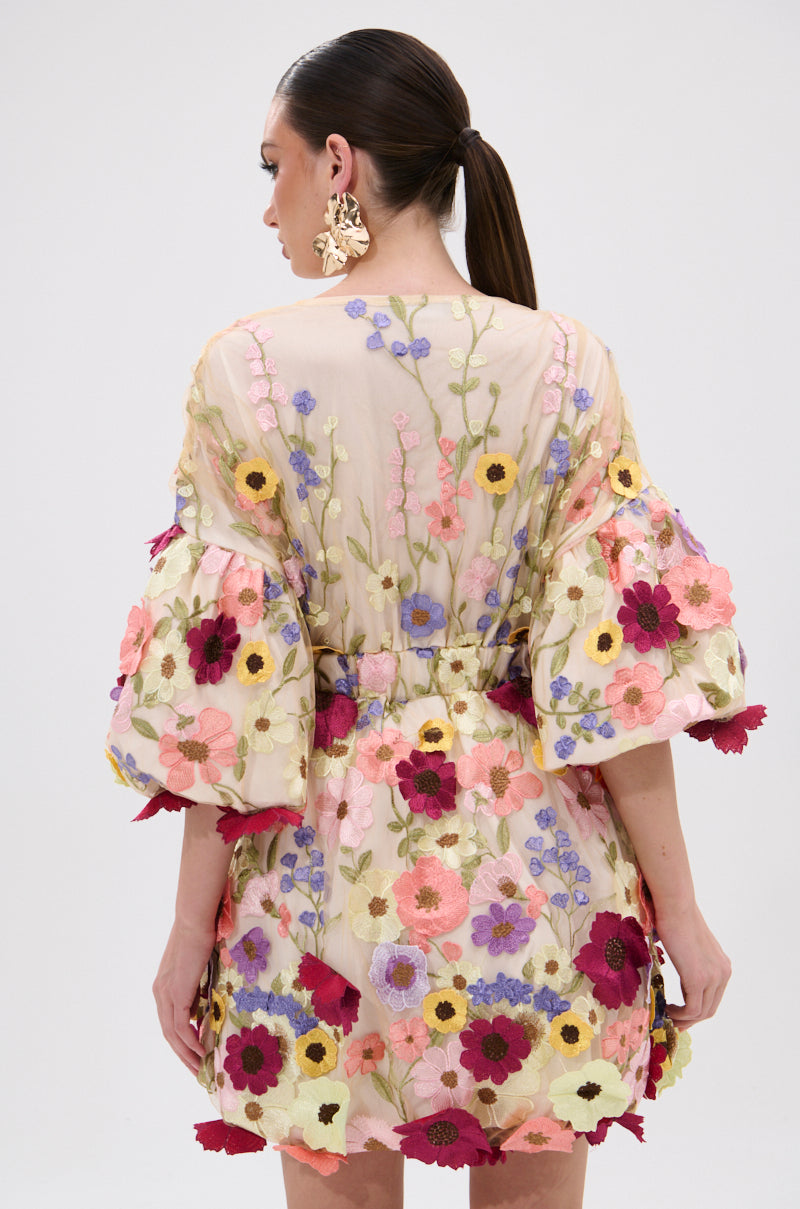A woman with dark hair in a low ponytail is seen from the back wearing the FLORAL BABY MINI DRESS, featuring colorful embroidered floral appliques, voluminous sleeves, and large floral earrings, against a plain white background. 4