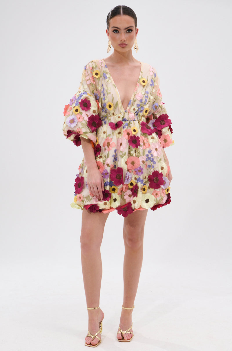 A woman wears the FLORAL BABY MINI DRESS, a floral mini with a short, deep V-neck and vibrant embroidered red, yellow, pink, and purple flower appliques. She styles it with slicked-back hair, gold earrings, and strappy sandals. 5
