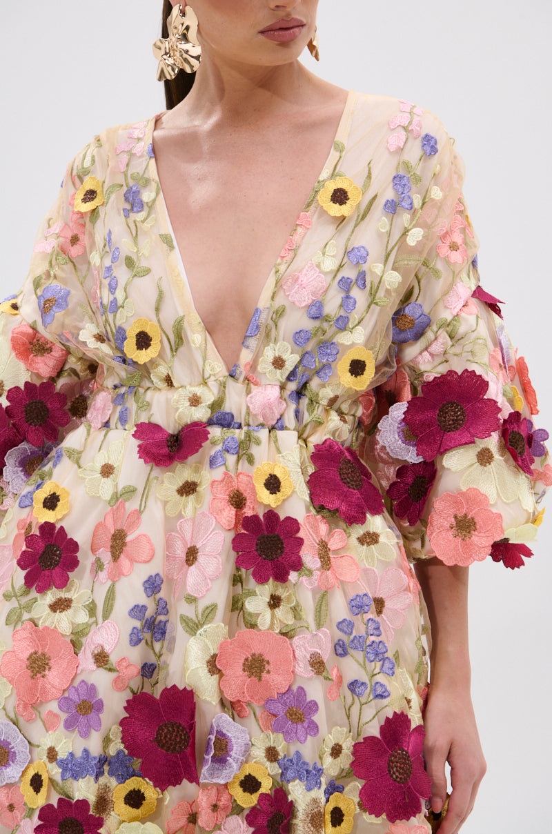 A woman wears the FLORAL BABY MINI DRESS, featuring a sheer design with a deep V-neckline and colorful embroidered flower appliques in pink, purple, yellow, and burgundy. She pairs it with statement floral earrings for a bold look. 7